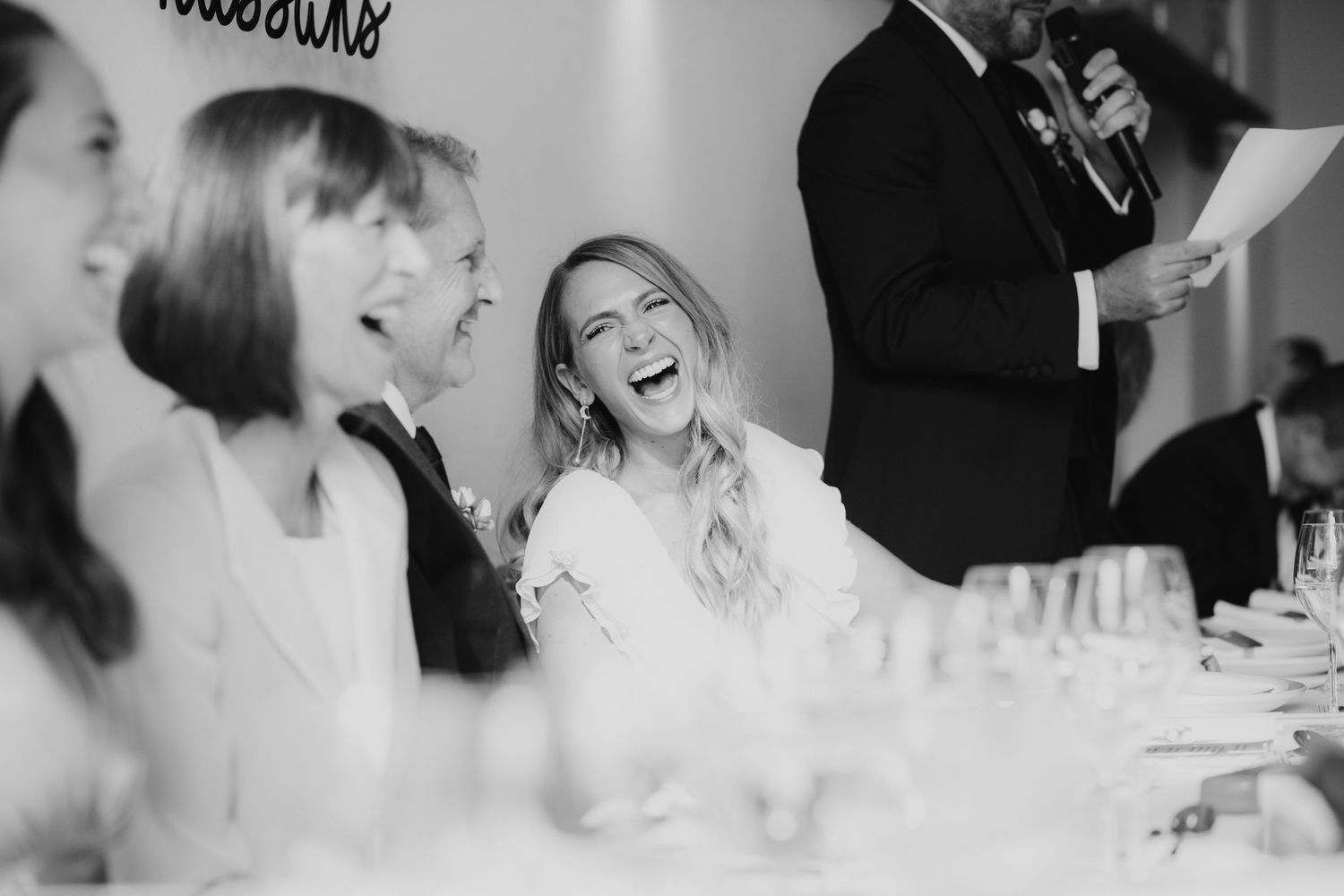 Black and white photo of bride laughing joyfully with guests during wedding reception speeches.