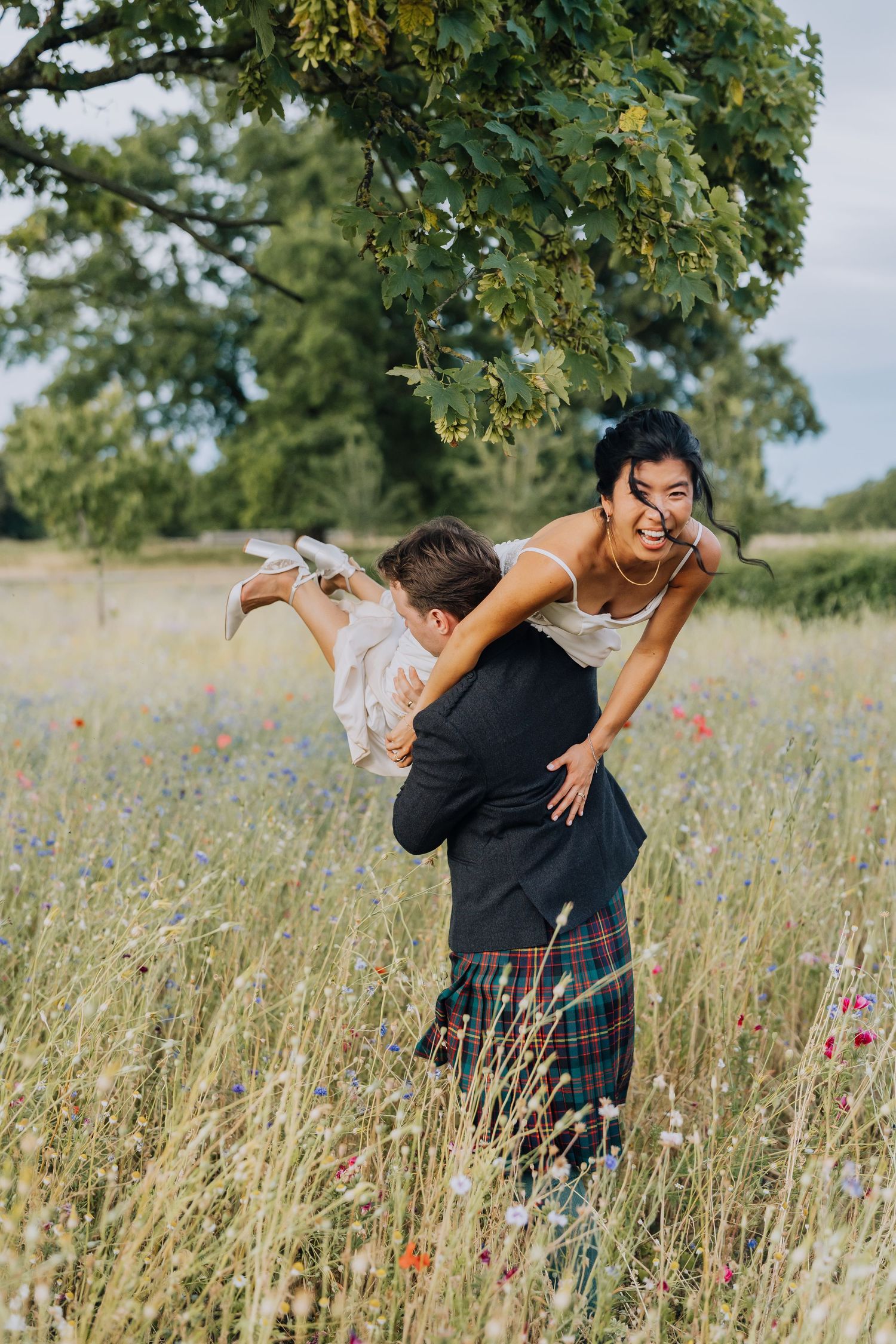 Couple playfully dancing in a wildflower meadow, groom in tartan kilt lifting laughing bride outdoors.