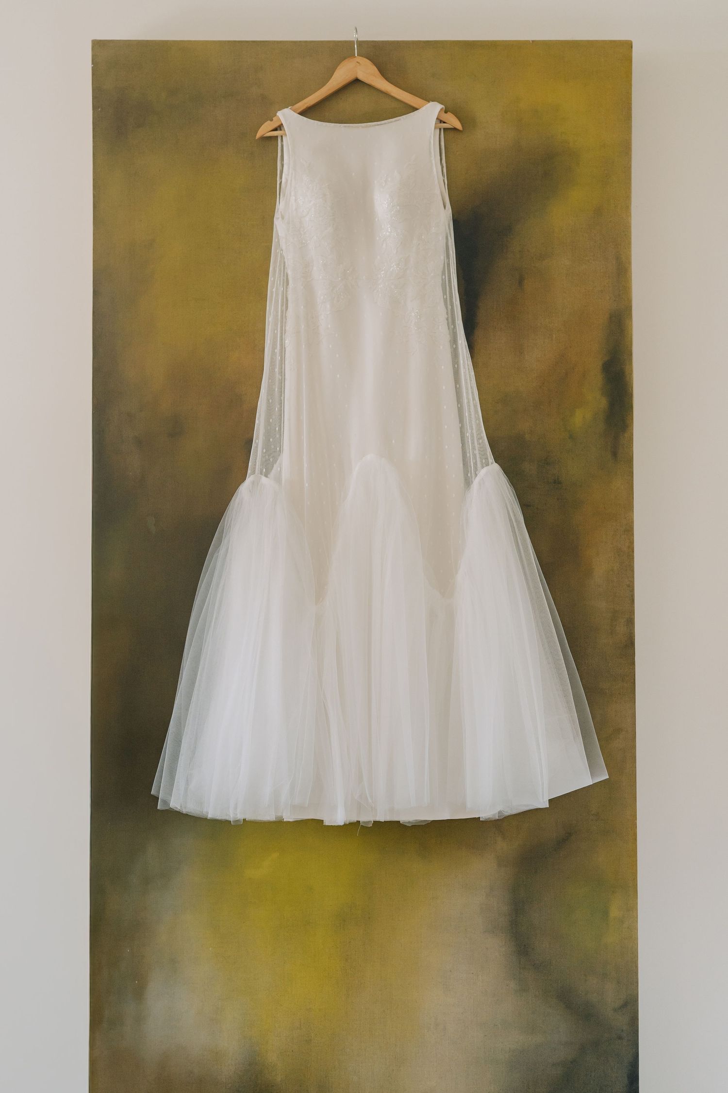 White sleeveless tulle wedding dress hanging on wooden hanger against abstract olive green painted canvas backdrop.
