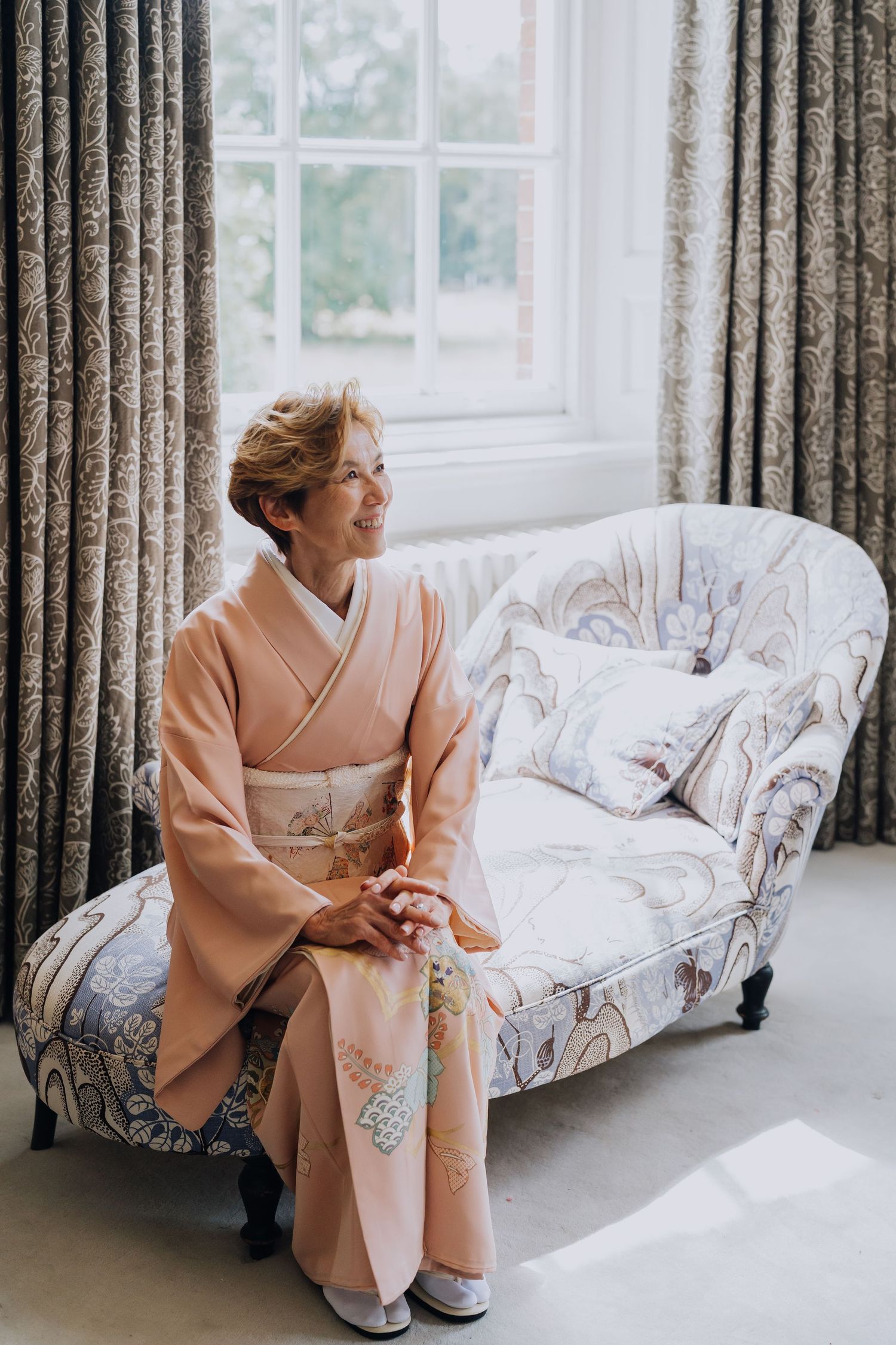 Elegant older woman in a blush pink kimono sitting gracefully on a floral chaise lounge by a sunlit window.