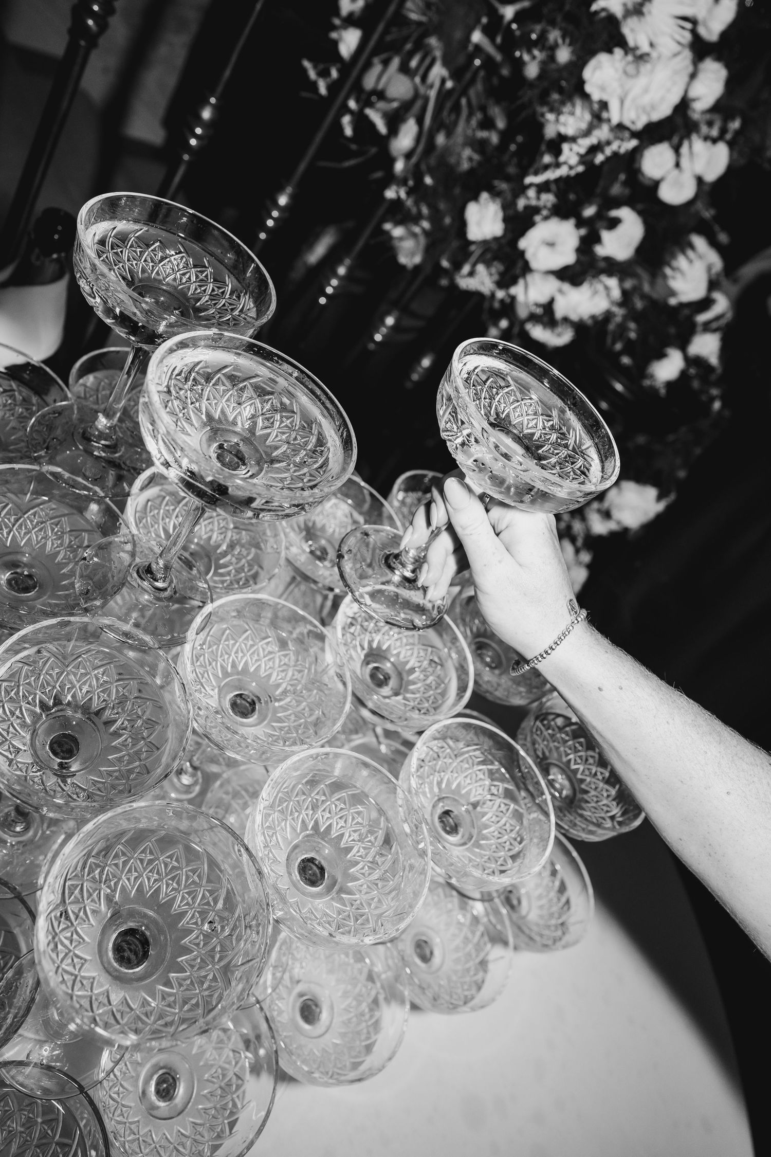 Black and white photo of hand reaching for crystal champagne tower at wedding reception.