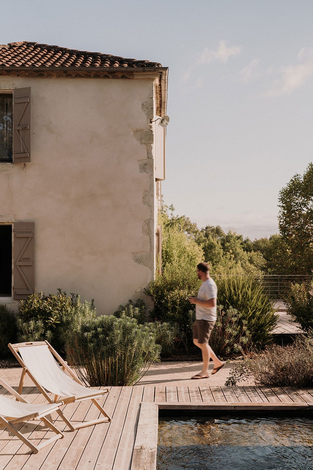 Man walking on wooden deck beside rustic French farmhouse with pool and lush Mediterranean garden.