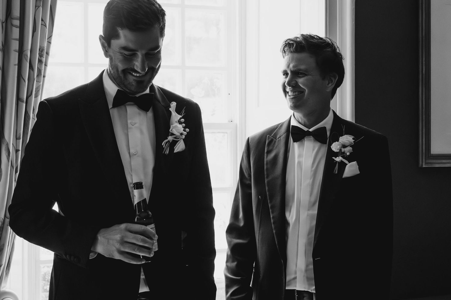 Two grooms in tuxedos with boutonnieres share a joyful moment in black and white wedding photo.