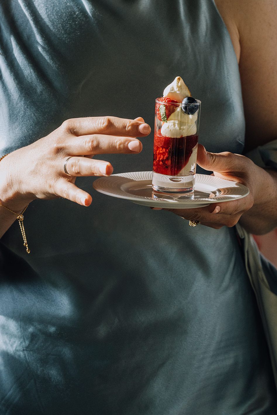Person in teal dress holding silver plate with layered berry and cream dessert shot glass.