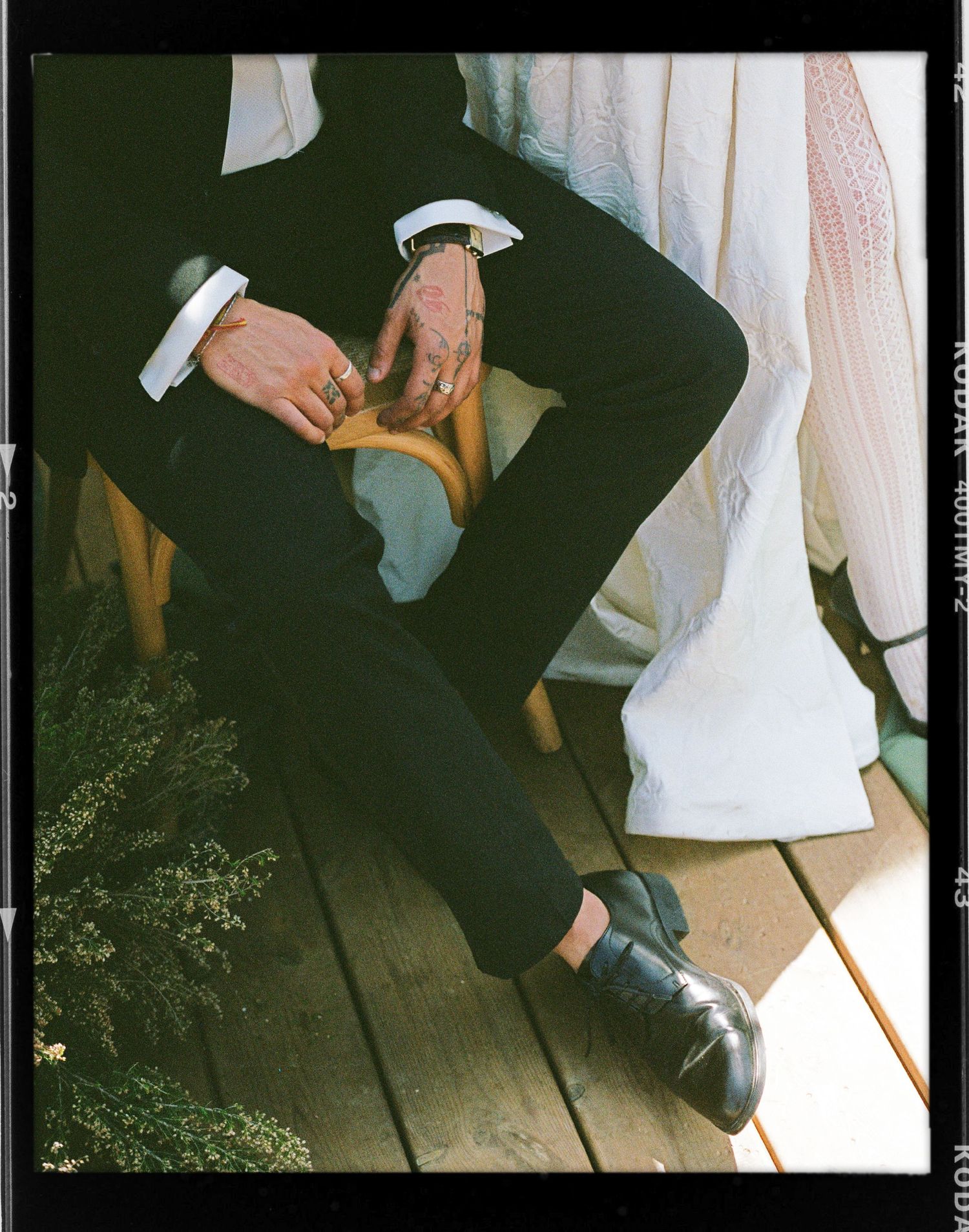 Groom in black suit with tattooed hands holding a wooden clutch, seated beside a white wedding dress on a sunlit deck.