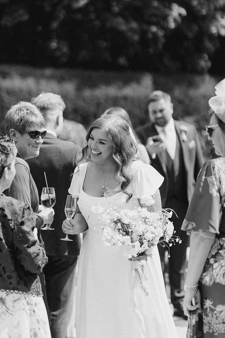 Smiling bride in white gown holding bouquet mingles with guests at outdoor wedding celebration.
