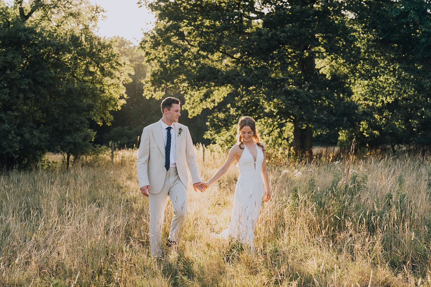 Bride and groom hold hands walking through golden sunlit meadow in romantic outdoor wedding portrait.
