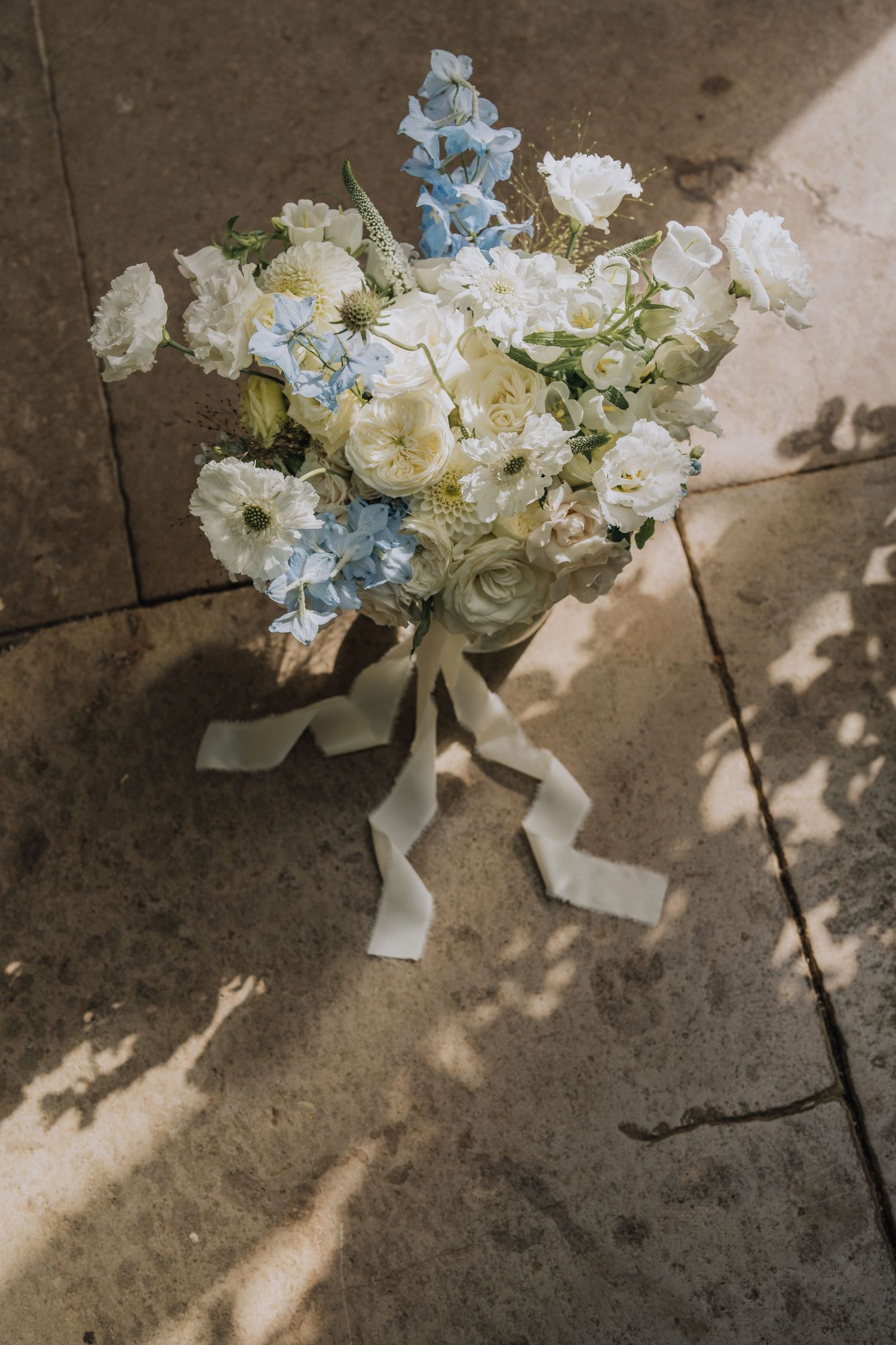 Romantic bridal bouquet of white roses and blue delphiniums with silk ribbon on stone floor.
