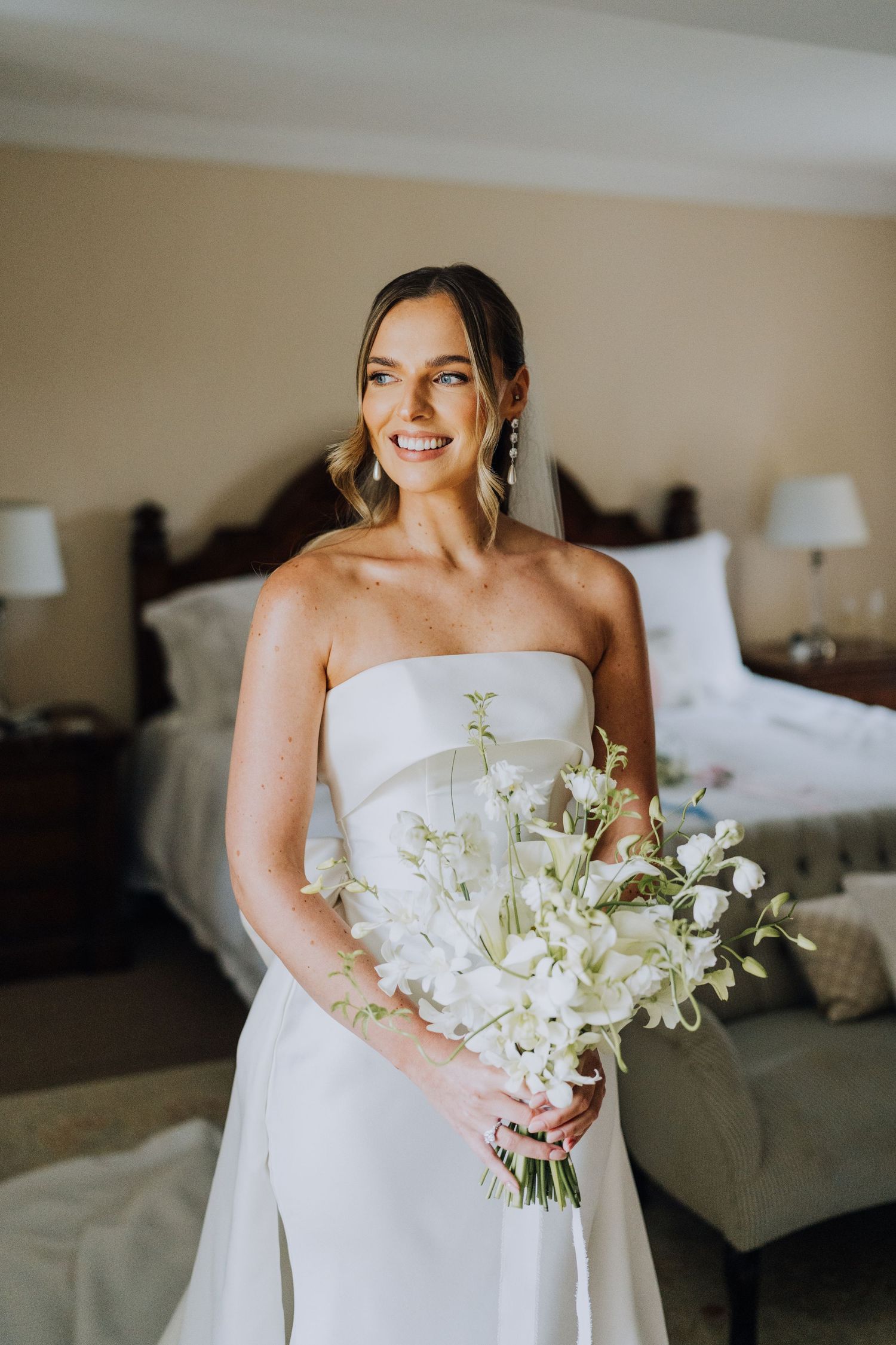 Smiling bride in strapless white gown holds elegant white floral bouquet in hotel room.