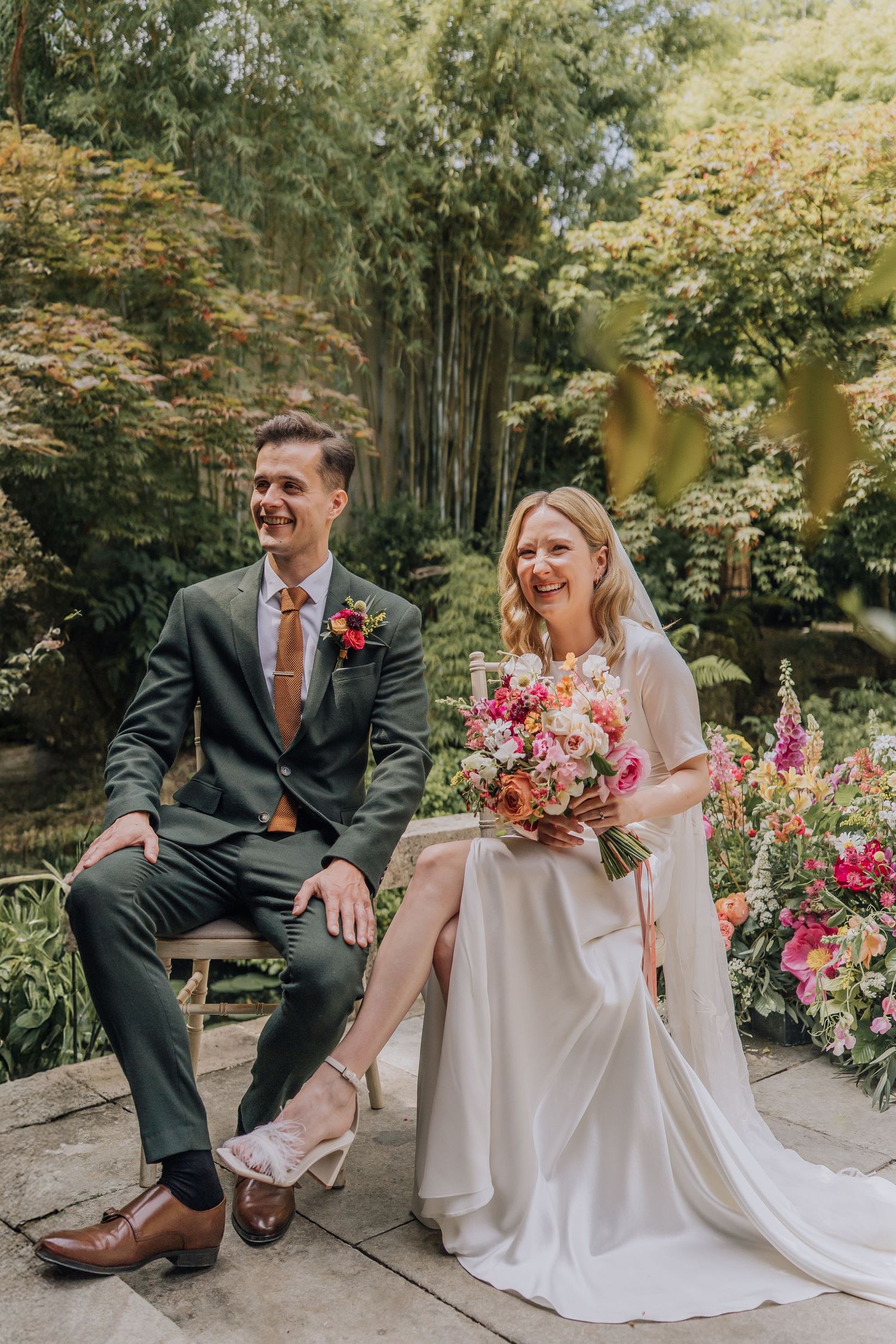 Laughing bride and groom sit together in lush garden surrounded by colorful summer flowers.