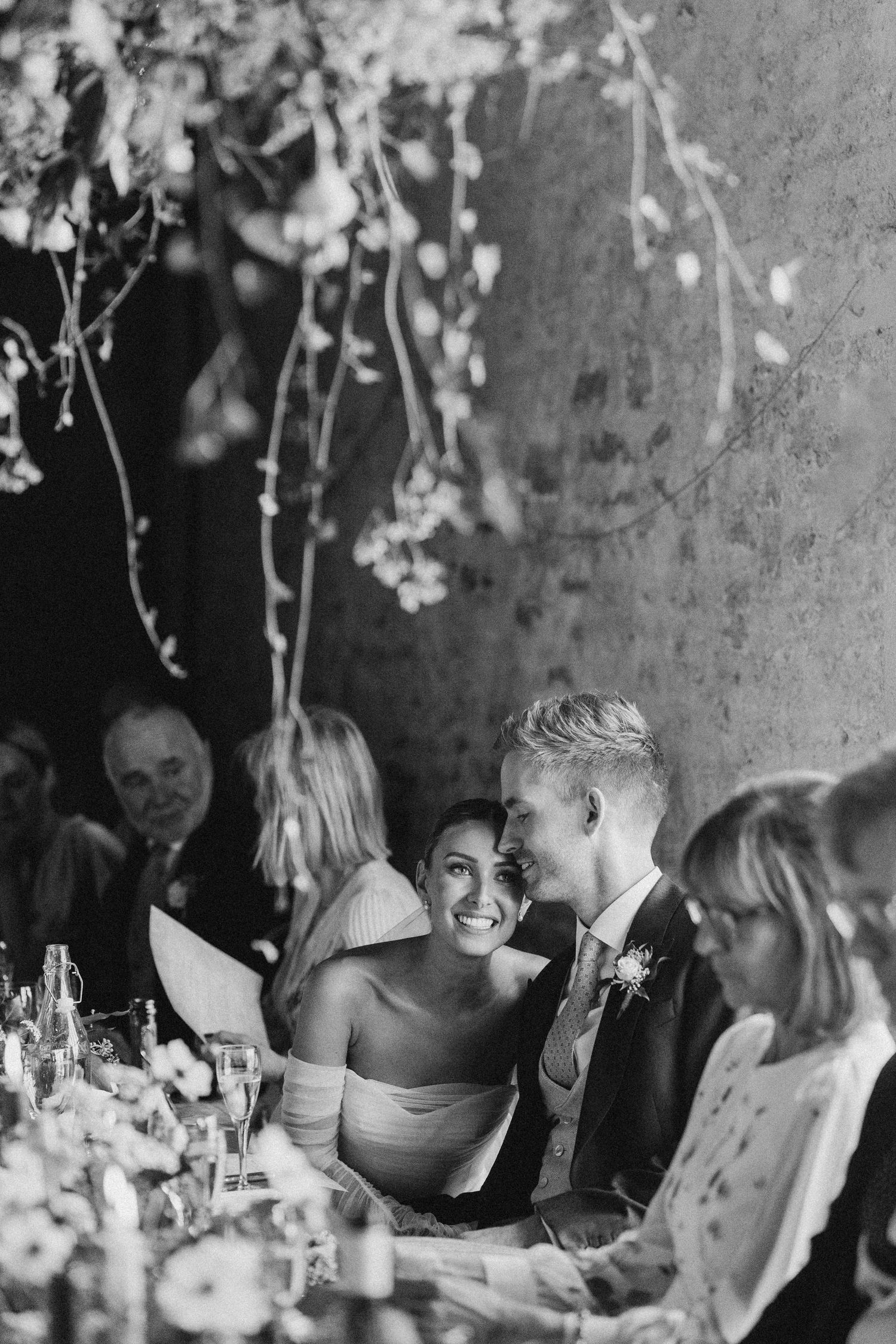 Black and white photo of bride and groom laughing at wedding reception table with floral decor.
