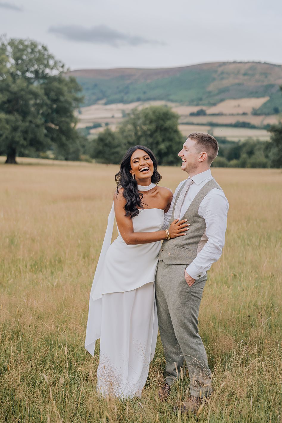 Laughing bride and groom embrace in golden meadow with rolling hills and countryside scenery behind them.