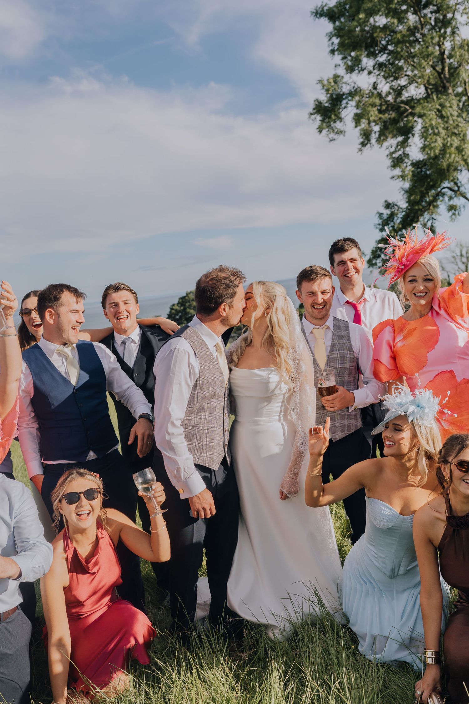 Bride and groom share a kiss surrounded by smiling wedding guests outdoors on a sunny day.