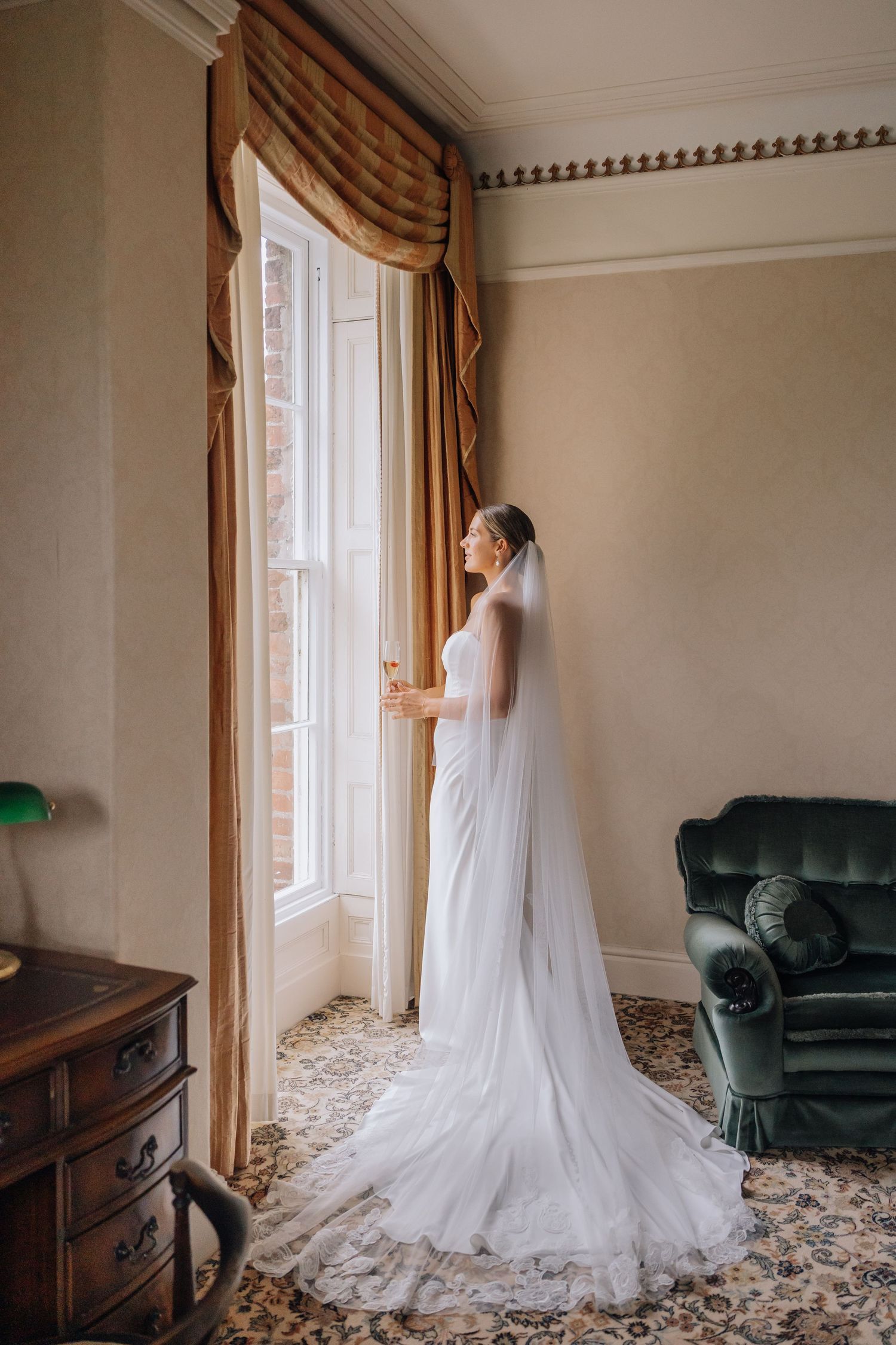 Bride in white lace gown and veil stands by tall window in elegant room with gold drapes and green armchair.