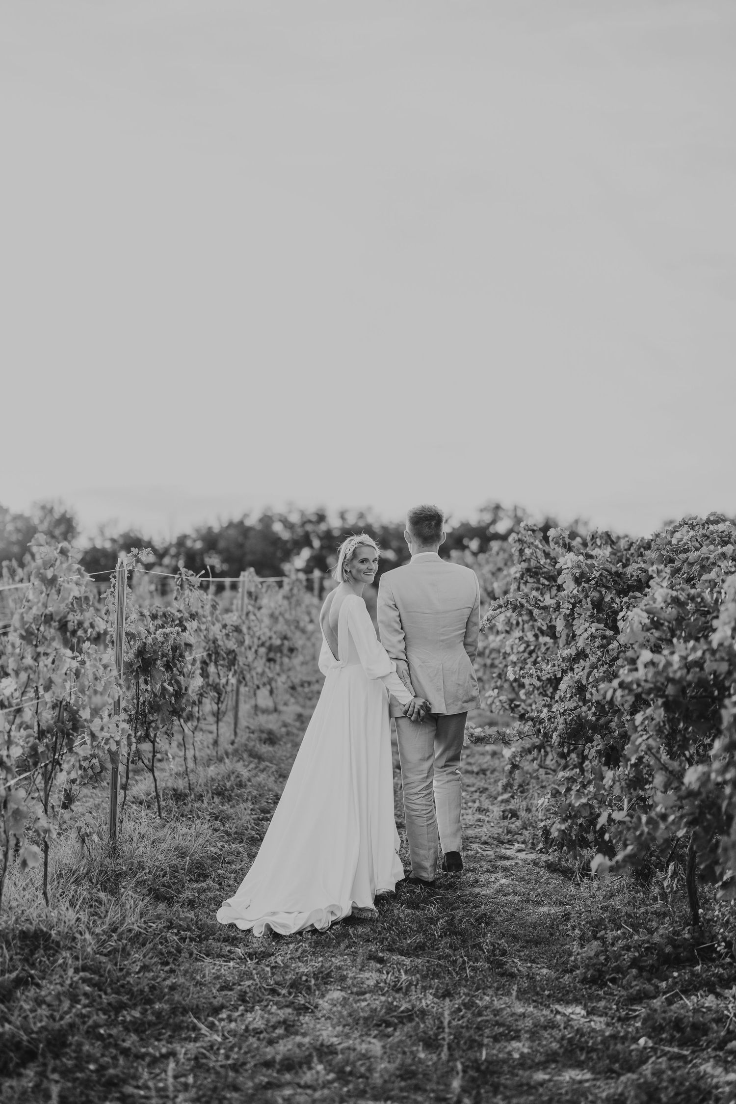 Black and white photo of bride and groom walking hand in hand through a lush vineyard on their wedding day.