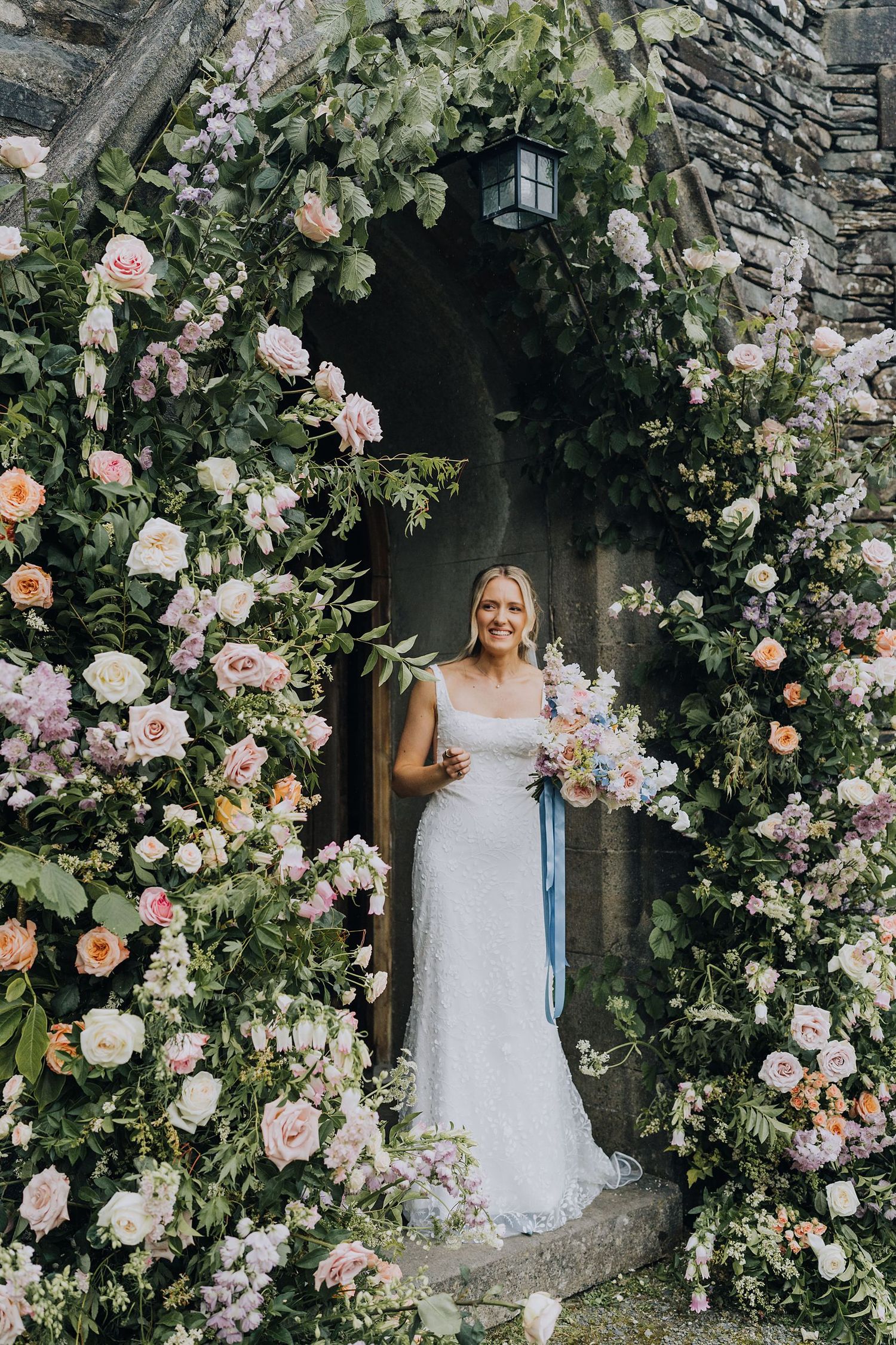 Bride in lace gown stands under a stunning floral arch of pink and white roses at a stone church.