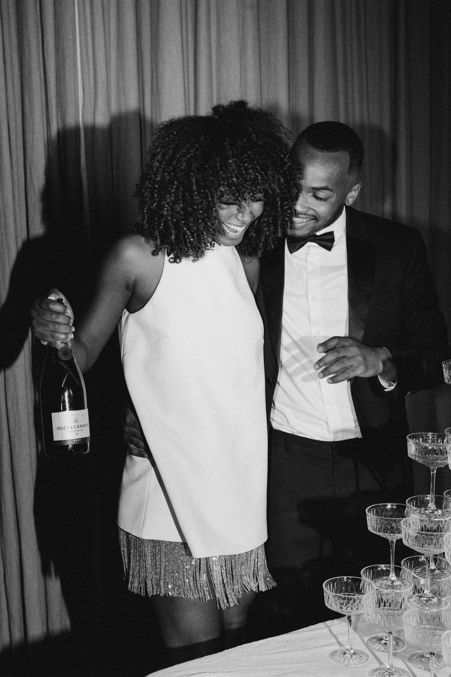 Joyful bride in white fringe mini dress and groom in tuxedo pose beside champagne tower at reception.