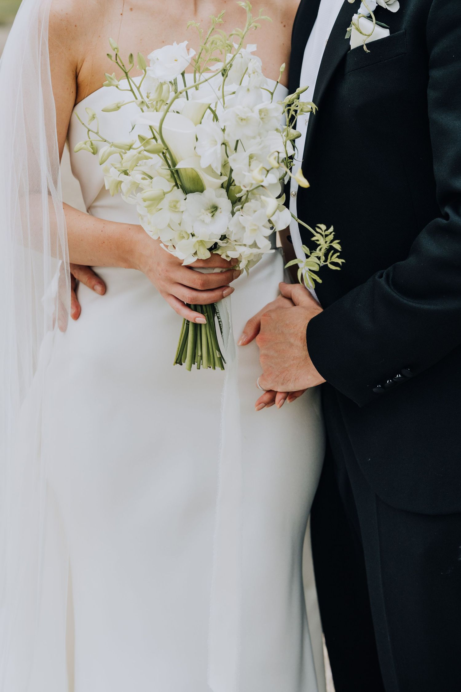 Bride and groom standing together, bride holding elegant white floral bouquet in fitted wedding gown.