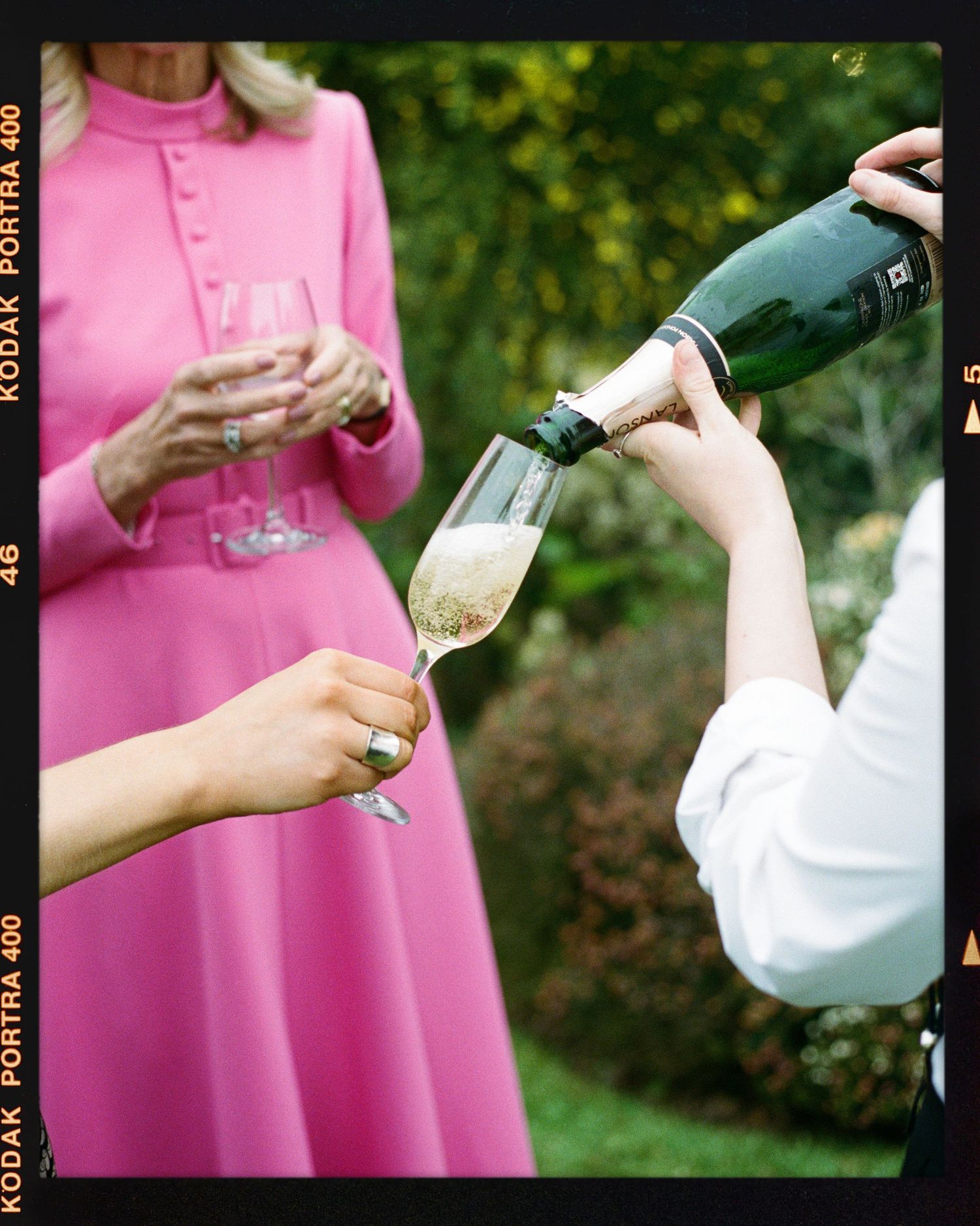 Champagne being poured into a flute at an outdoor garden party, with a woman in a pink dress holding a glass.