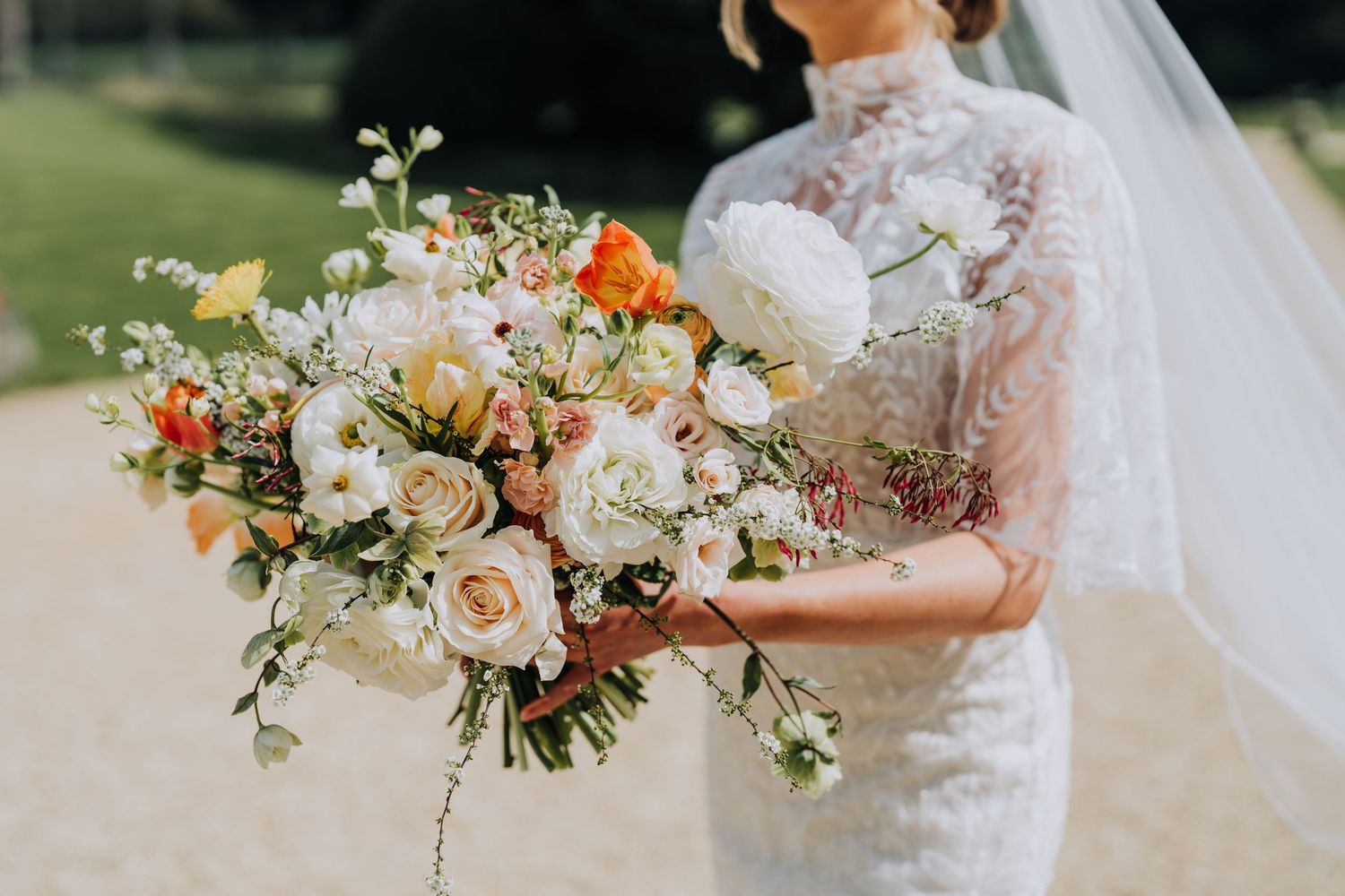 Bride holding a lush wedding bouquet with white, peach, and orange blooms in a lace gown outdoors.