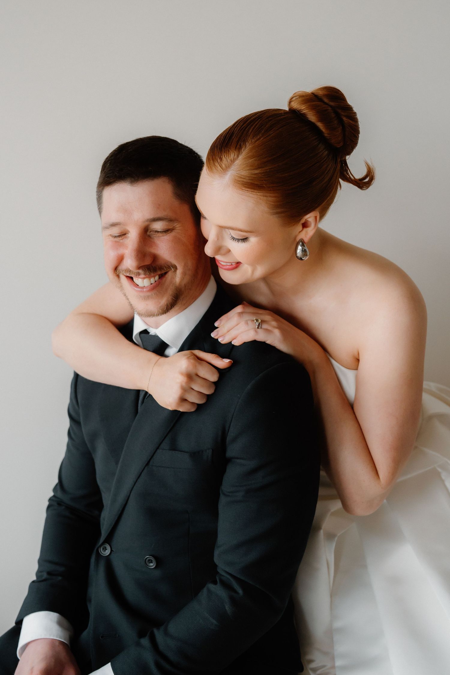 A smiling groom in a dark suit embraces a redheaded bride in a white strapless gown during their wedding portrait.