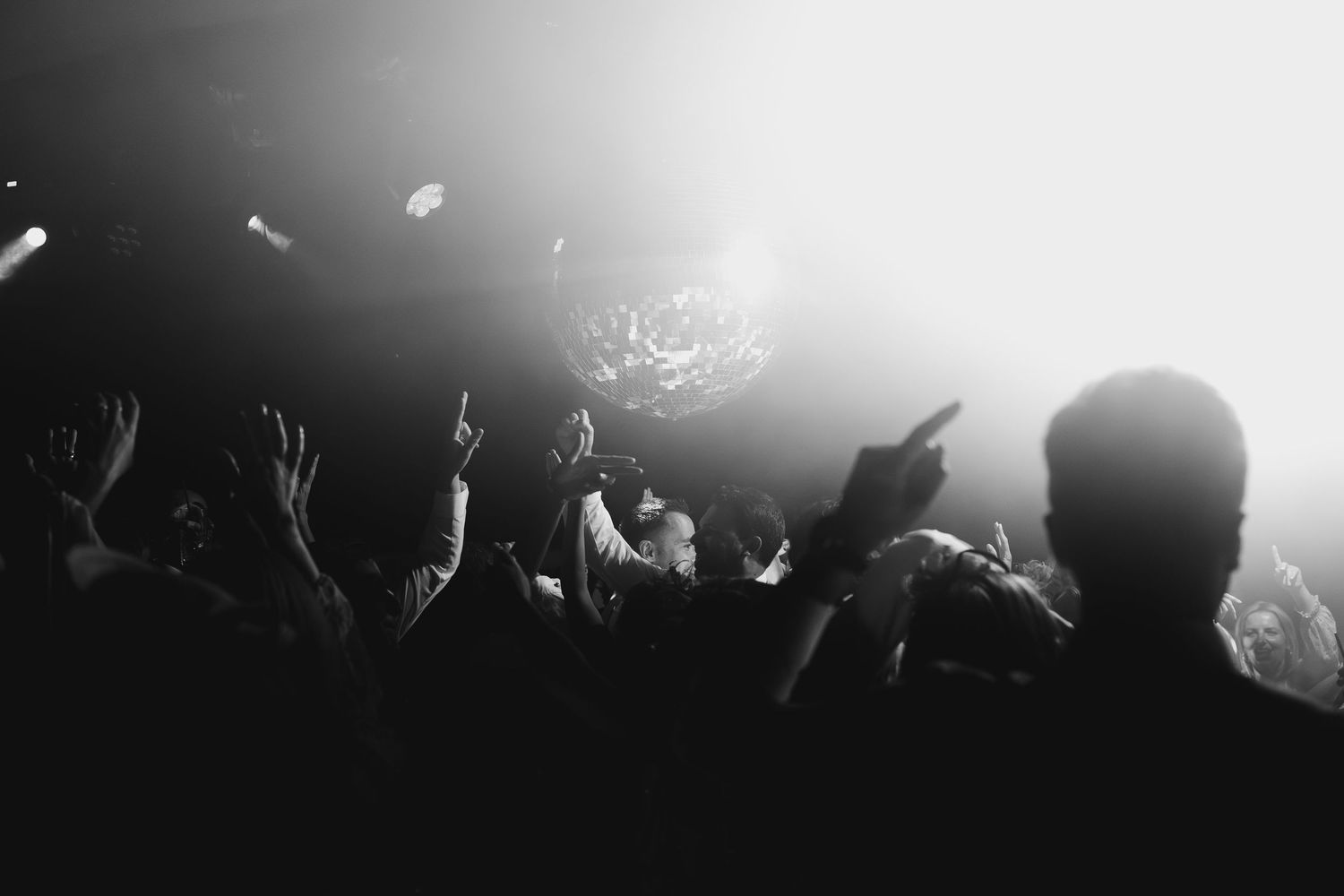 Black and white photo of concert crowd with hands raised under dramatic stage lighting.