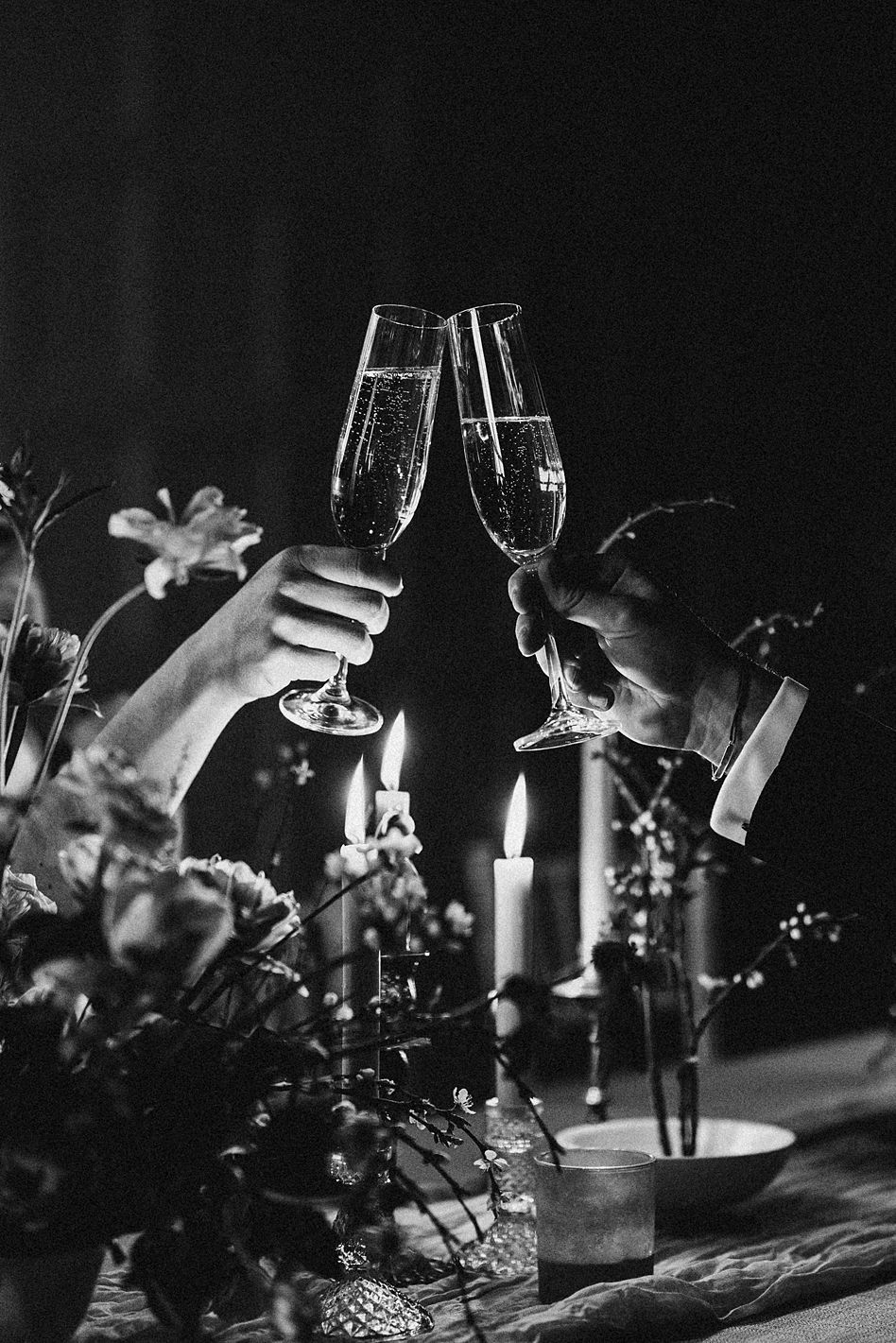 Black and white close-up of couple toasting champagne flutes by candlelight with floral arrangements at wedding reception.