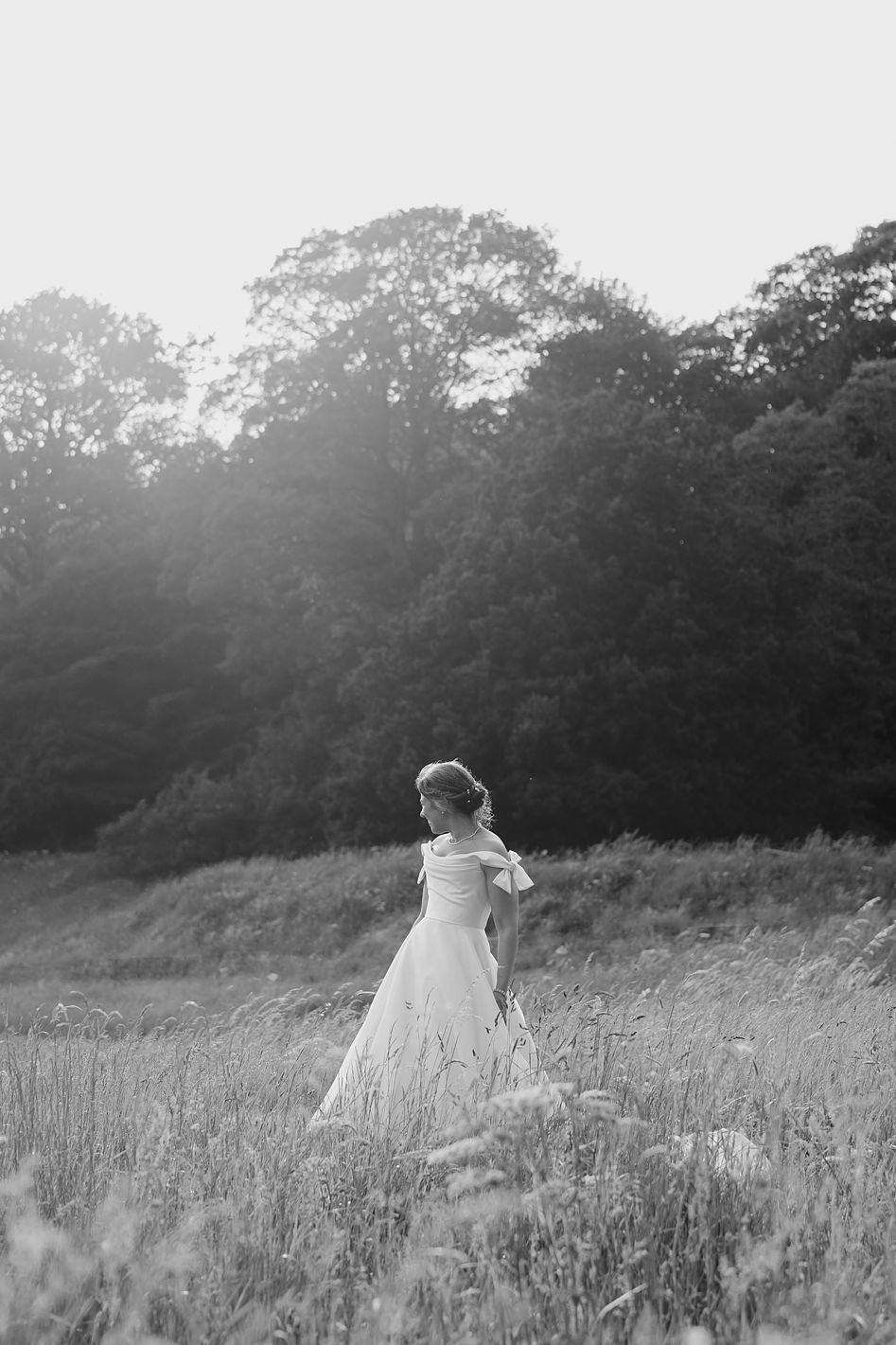 Black and white portrait of bride in off-shoulder ball gown standing alone in sunlit field at sunset.