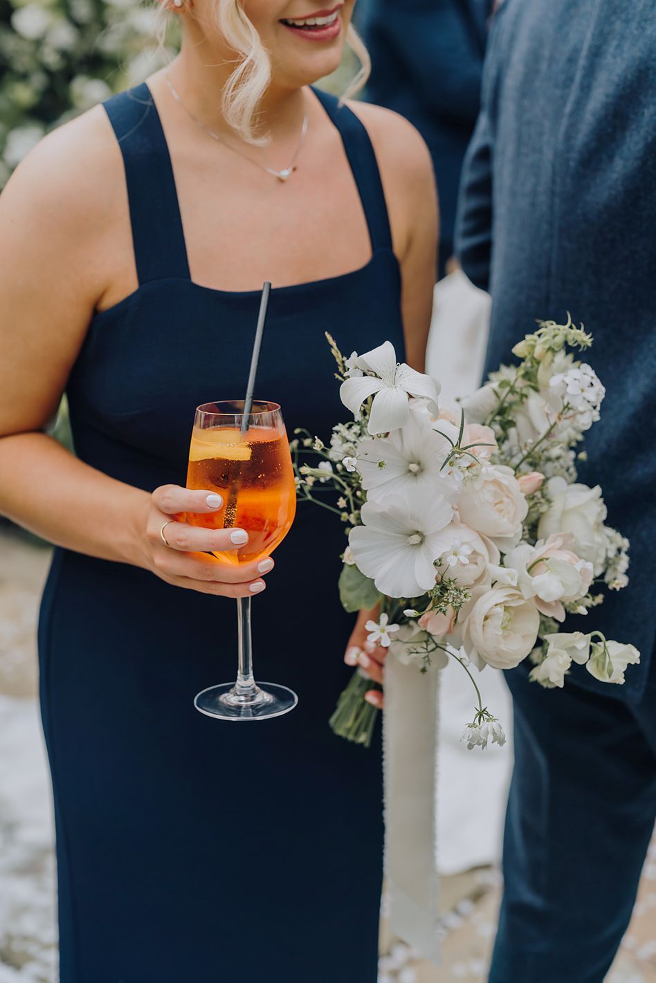 Bridesmaid in navy dress holding white floral bouquet and Aperol Spritz cocktail at wedding reception.
