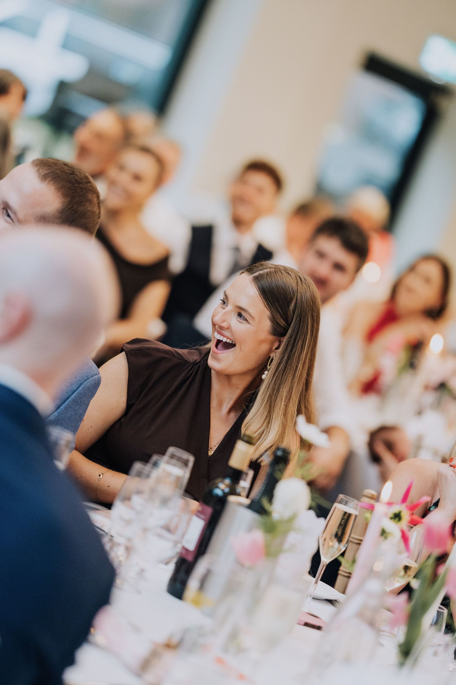 Laughing female wedding guest in dark dress enjoying speeches at an elegant indoor reception dinner.