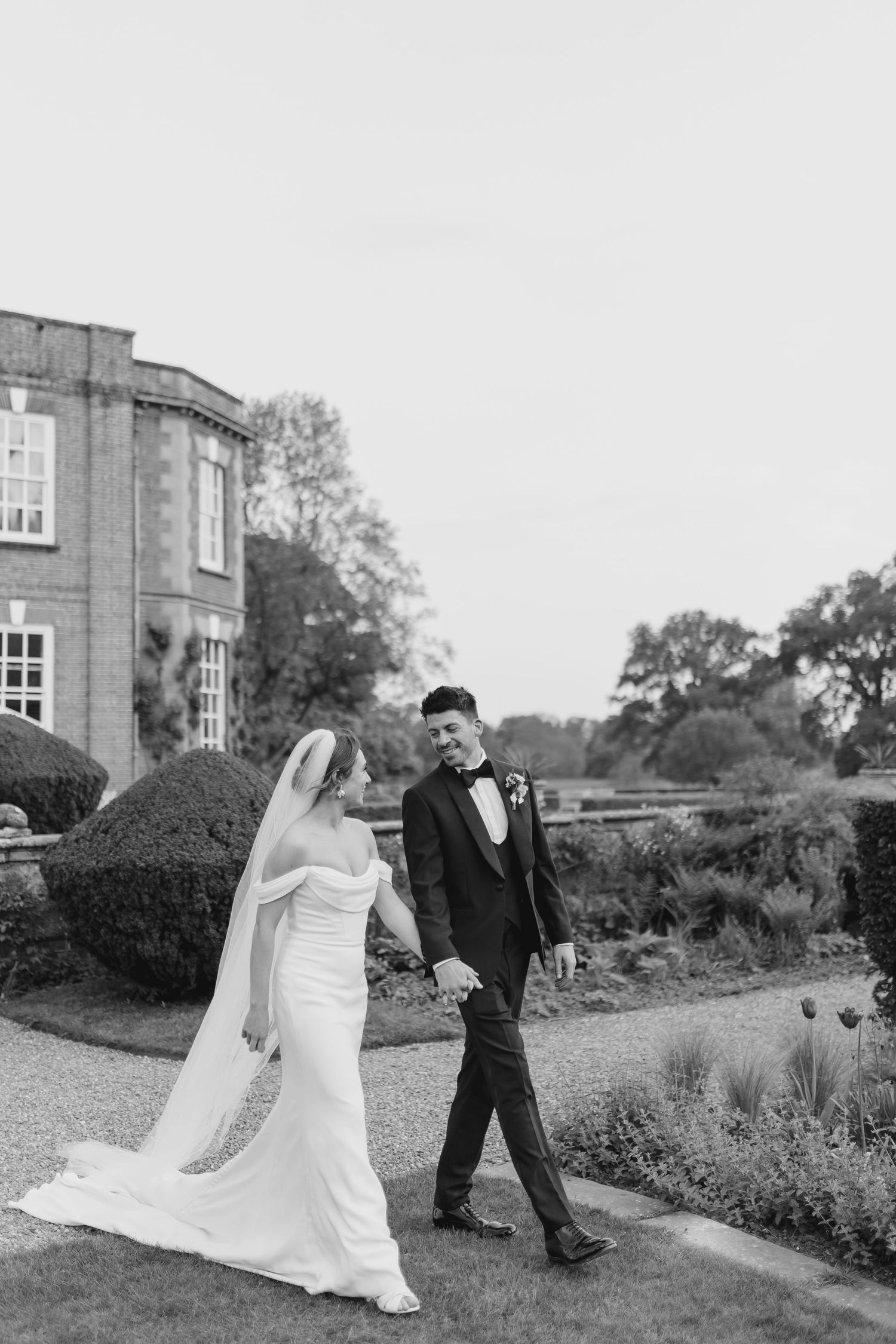 Black and white photo of bride and groom walking hand in hand in formal gardens of a stately manor house.