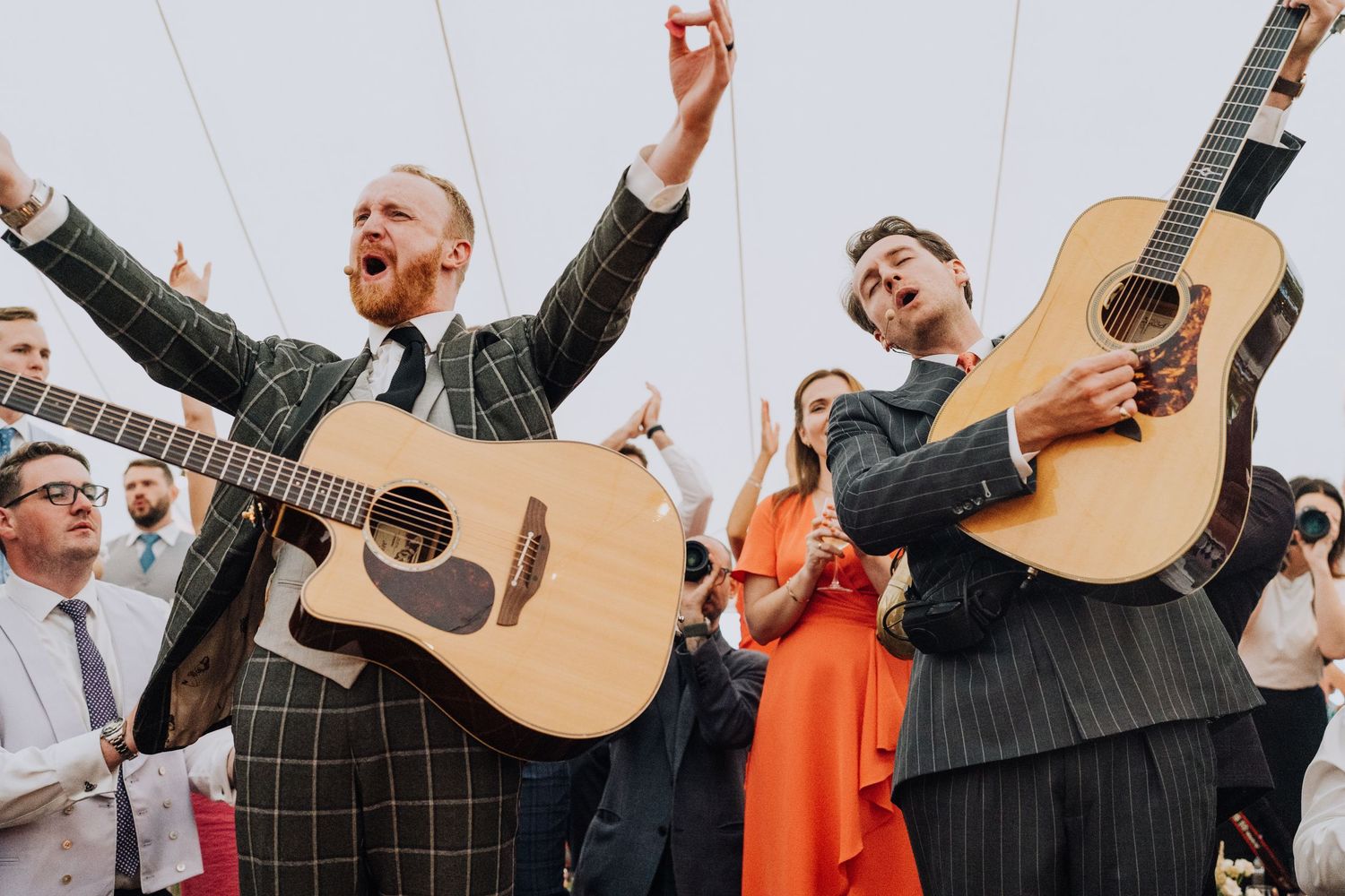 Energetic acoustic guitar duo performing at wedding reception, raising arms in celebration under tent.