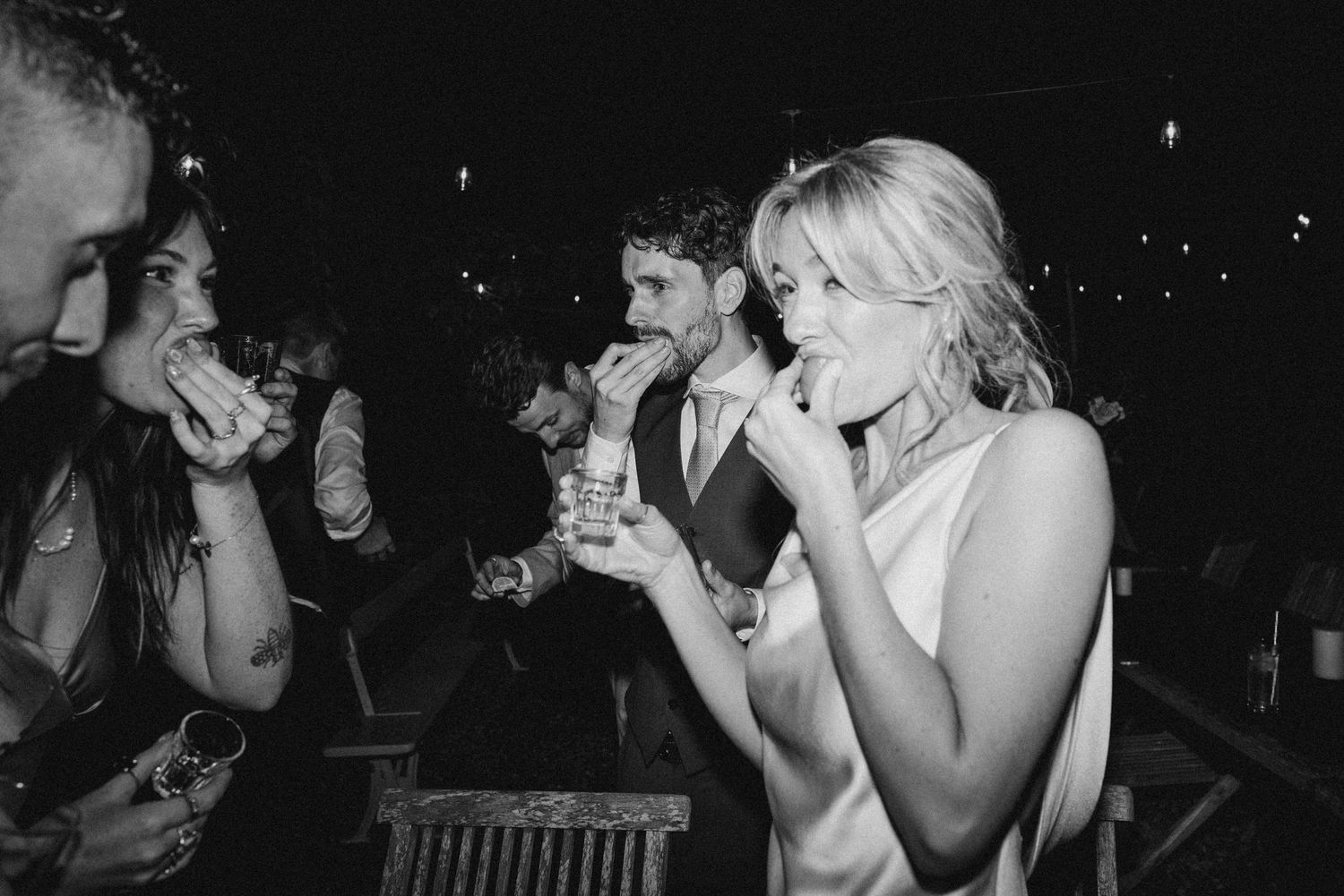 Black and white photo of wedding guests laughing and drinking at a lively evening reception party.