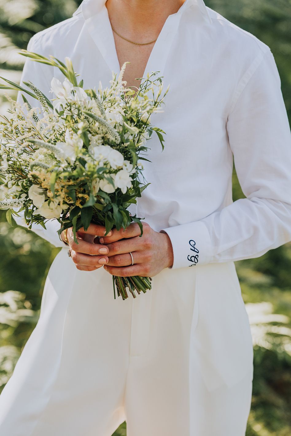 Bride in white pantsuit holds a delicate bouquet of white wildflowers with embroidered cuffs.