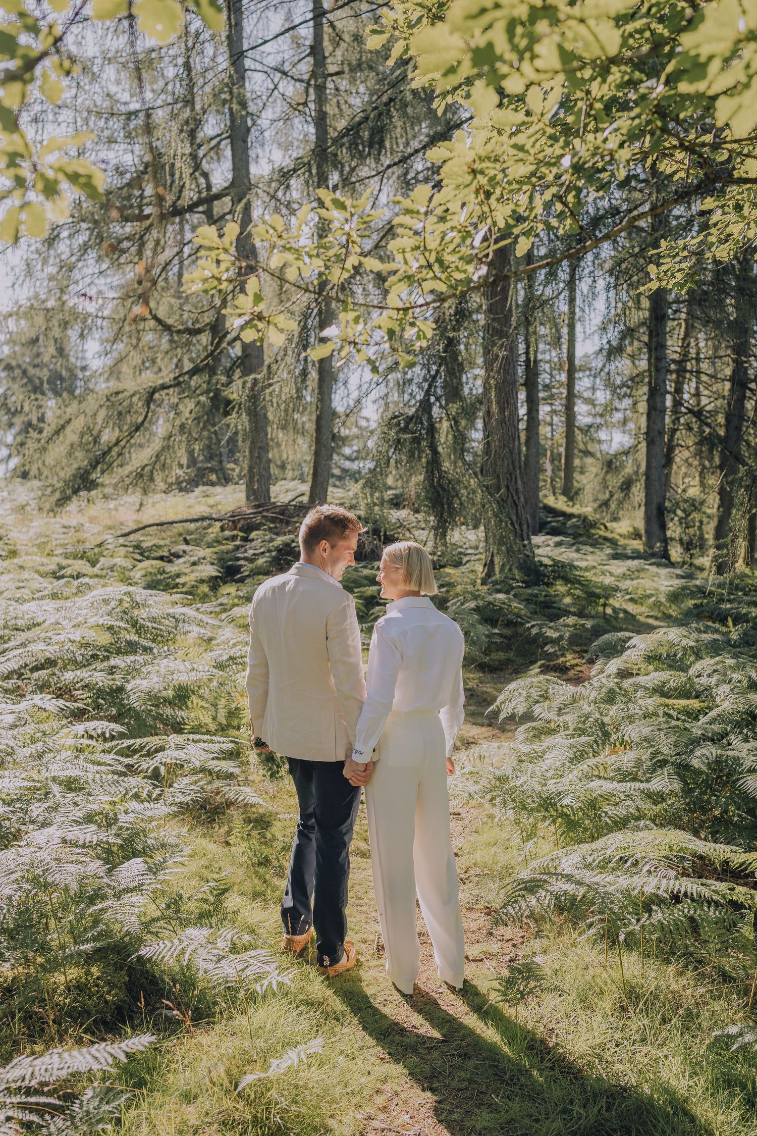 Wedding couple holding hands stroll along a forest trail lined with ferns and towering trees in golden sunlight.