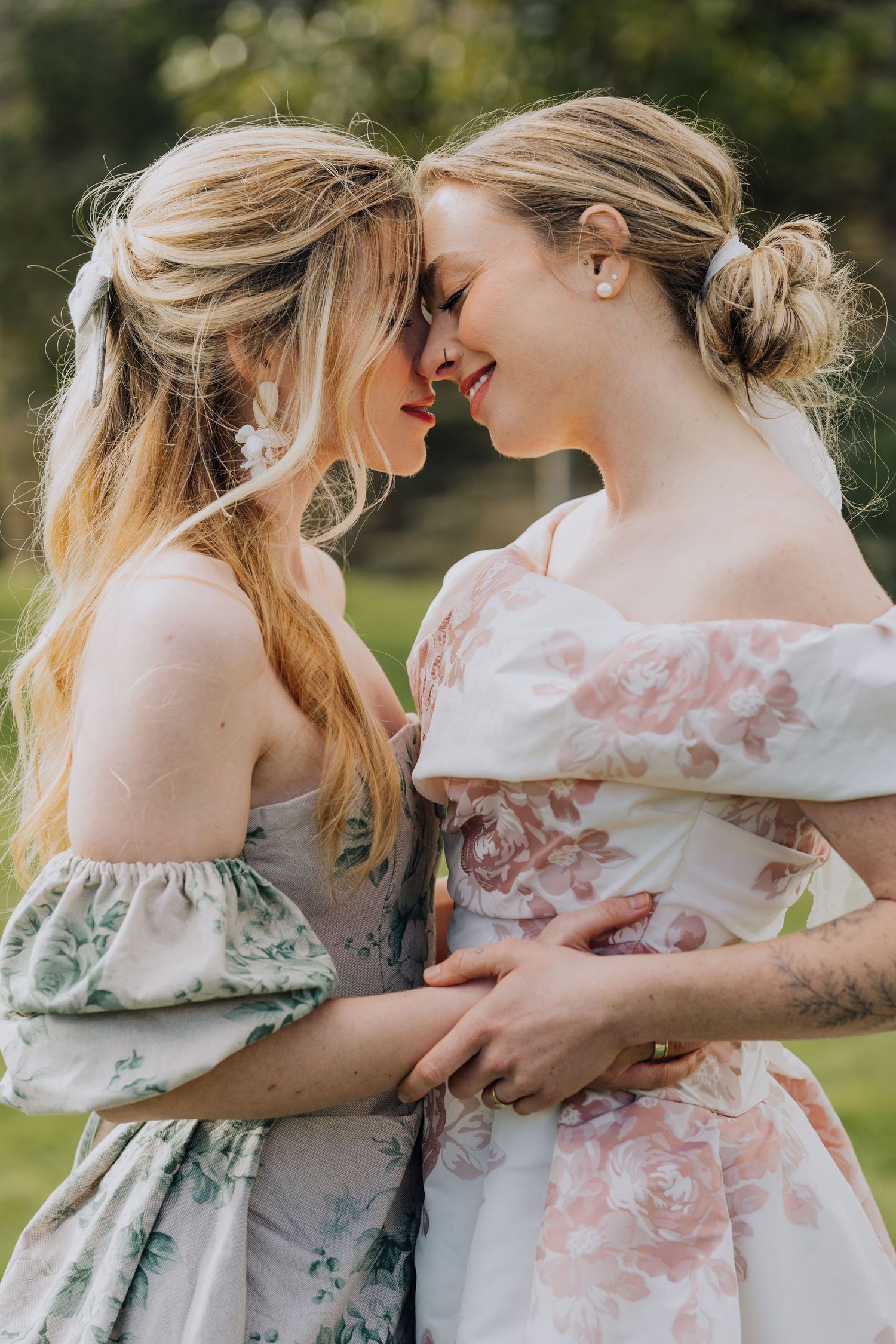 Two blonde women in floral off-shoulder dresses share a romantic moment outdoors, foreheads touching and smiling.