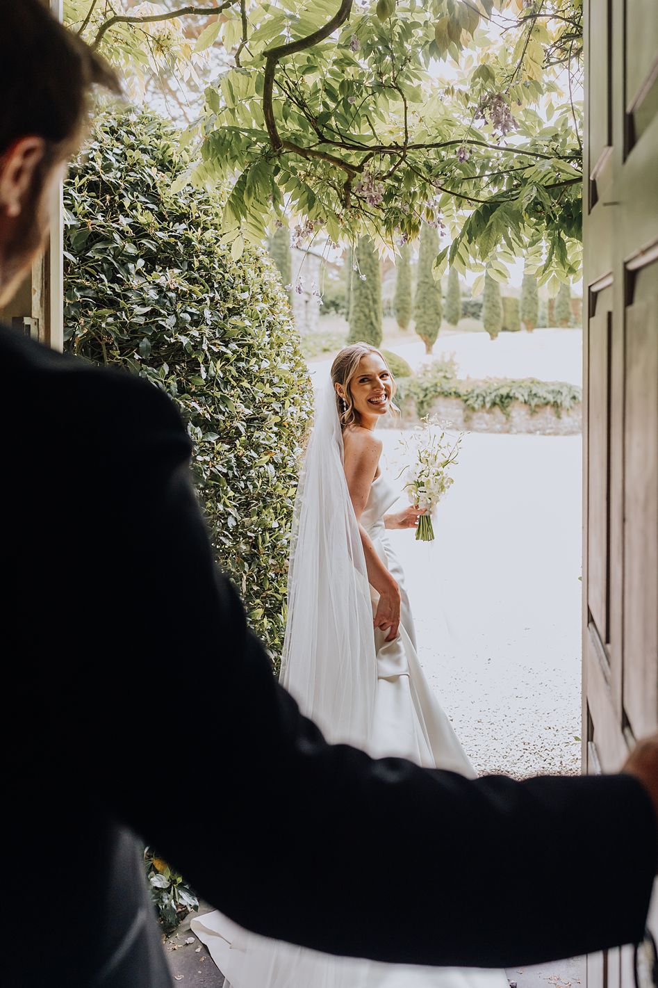 Smiling bride in a flowing white gown and veil stands in a garden doorway holding a bouquet.