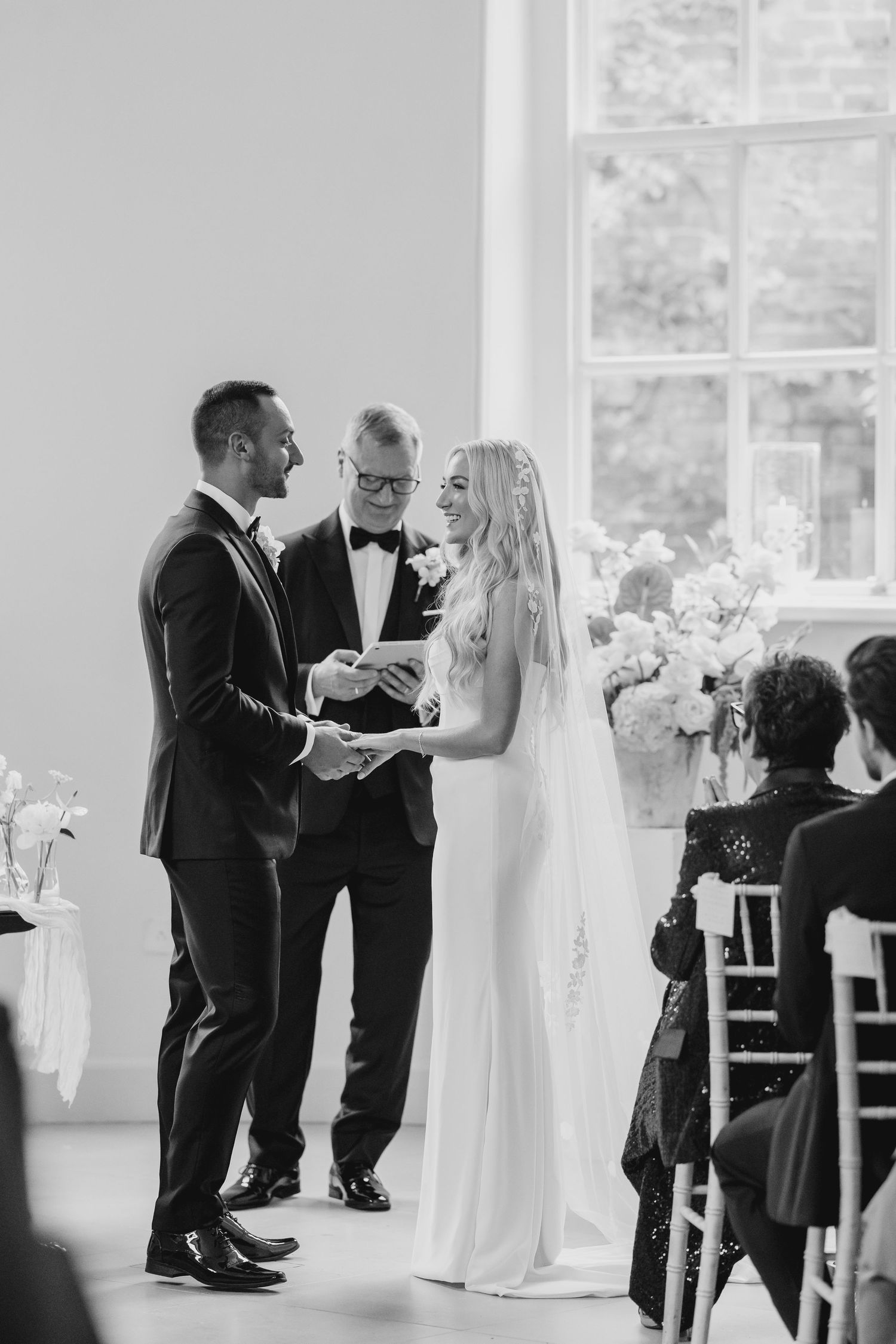 Black and white wedding ceremony photo of couple exchanging rings before officiant in bright elegant venue.