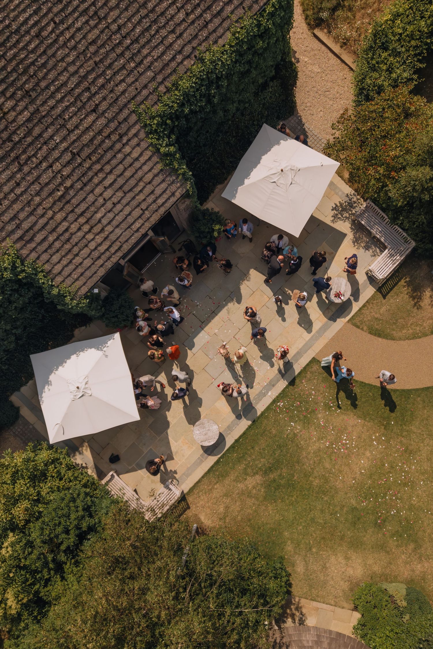 Aerial view of an outdoor patio event with guests mingling under white parasol umbrellas beside a stone building.