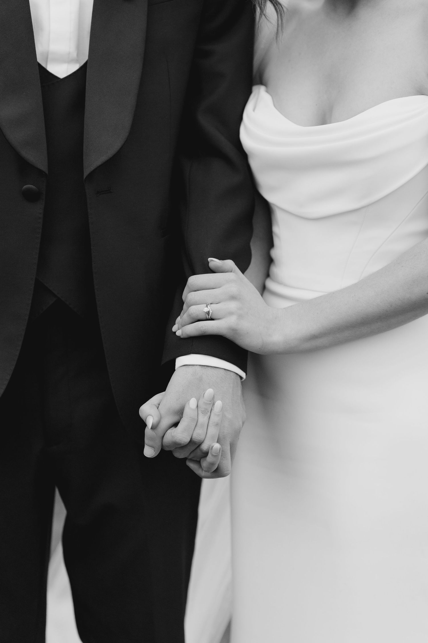 Black and white close-up of bride and groom holding hands, bride wearing diamond engagement ring in white gown.