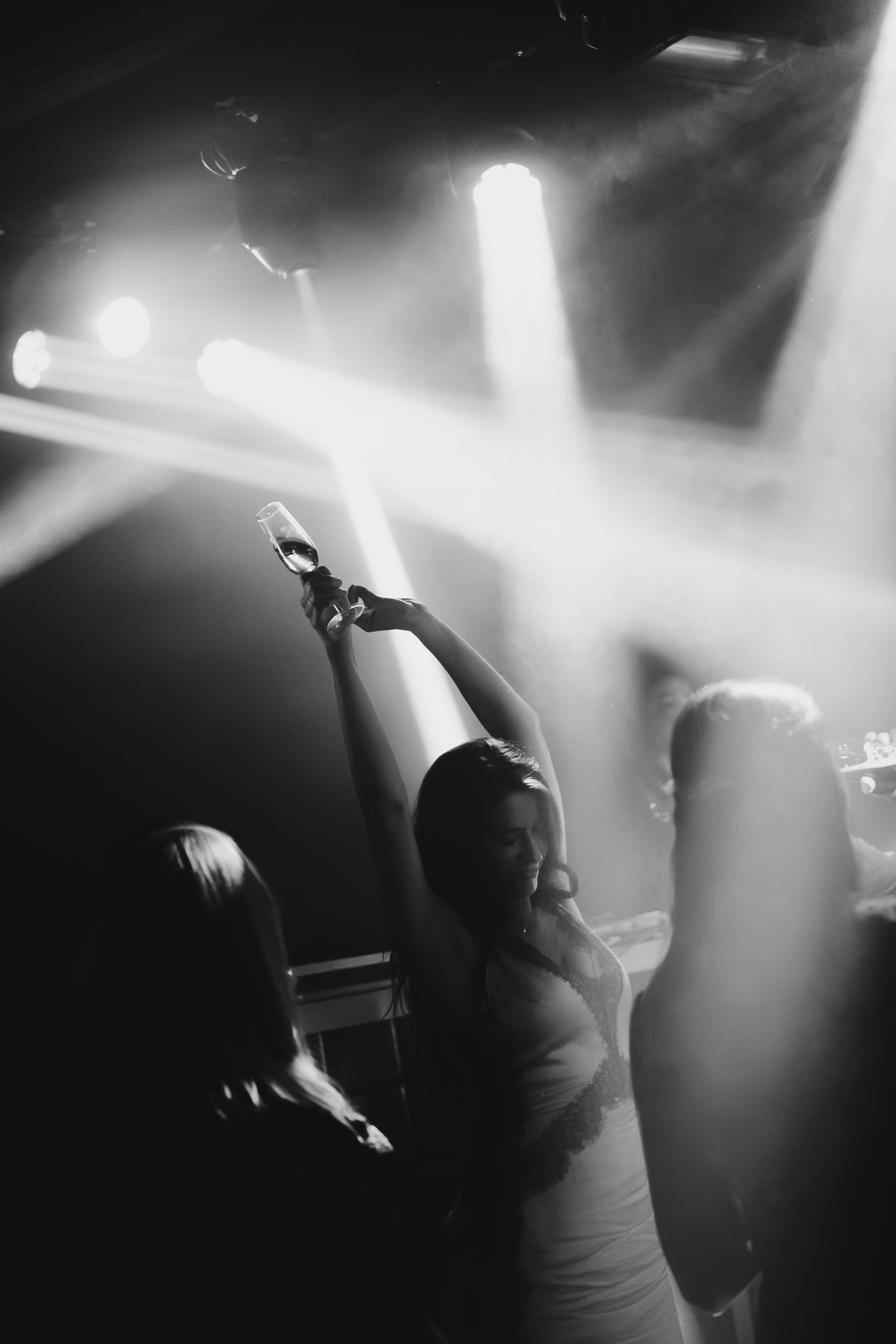Black and white photo of a bride dancing and raising a glass under dramatic stage lights at wedding reception.