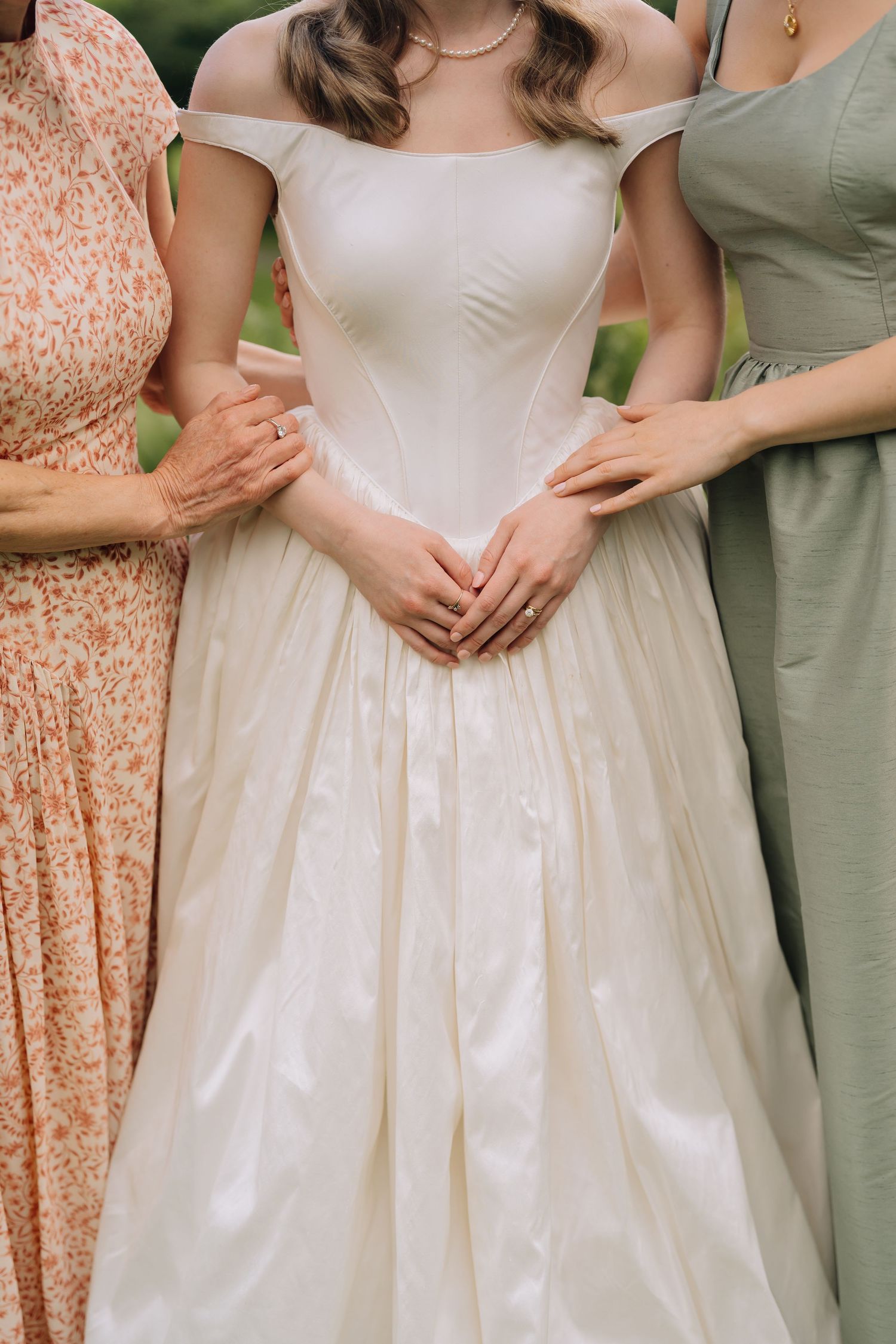 Bride in elegant off-shoulder satin ball gown flanked by two women in floral and sage dresses at outdoor wedding.