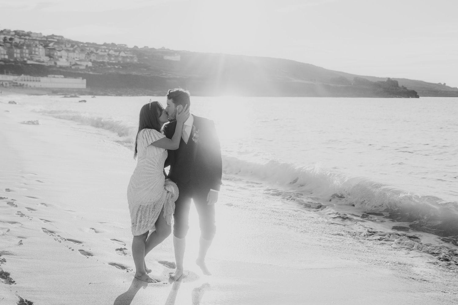 Black and white romantic photo of couple kissing on sunlit beach with ocean waves.