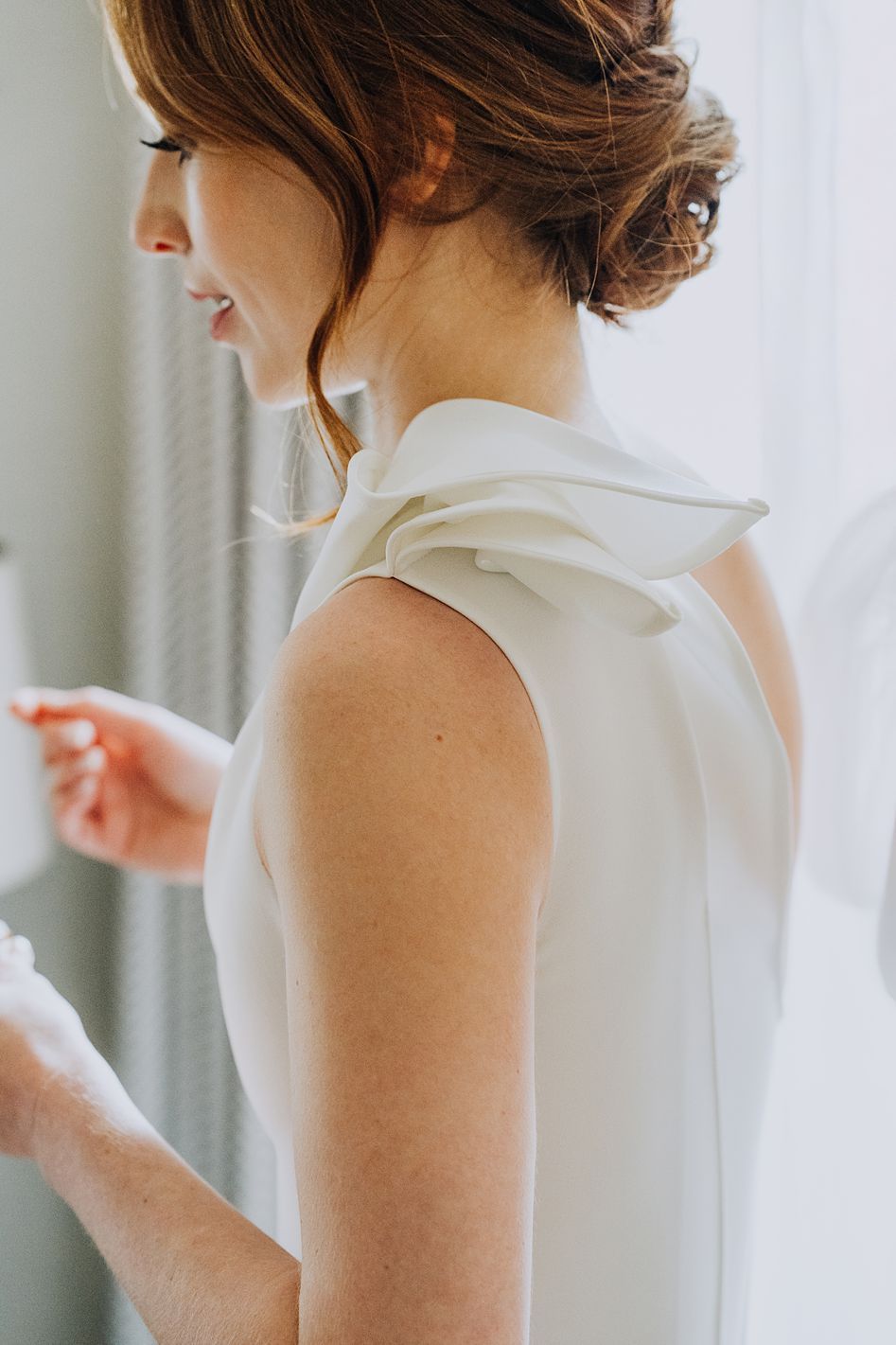Close-up of bride in elegant white gown with ruffle shoulder detail being helped to dress.