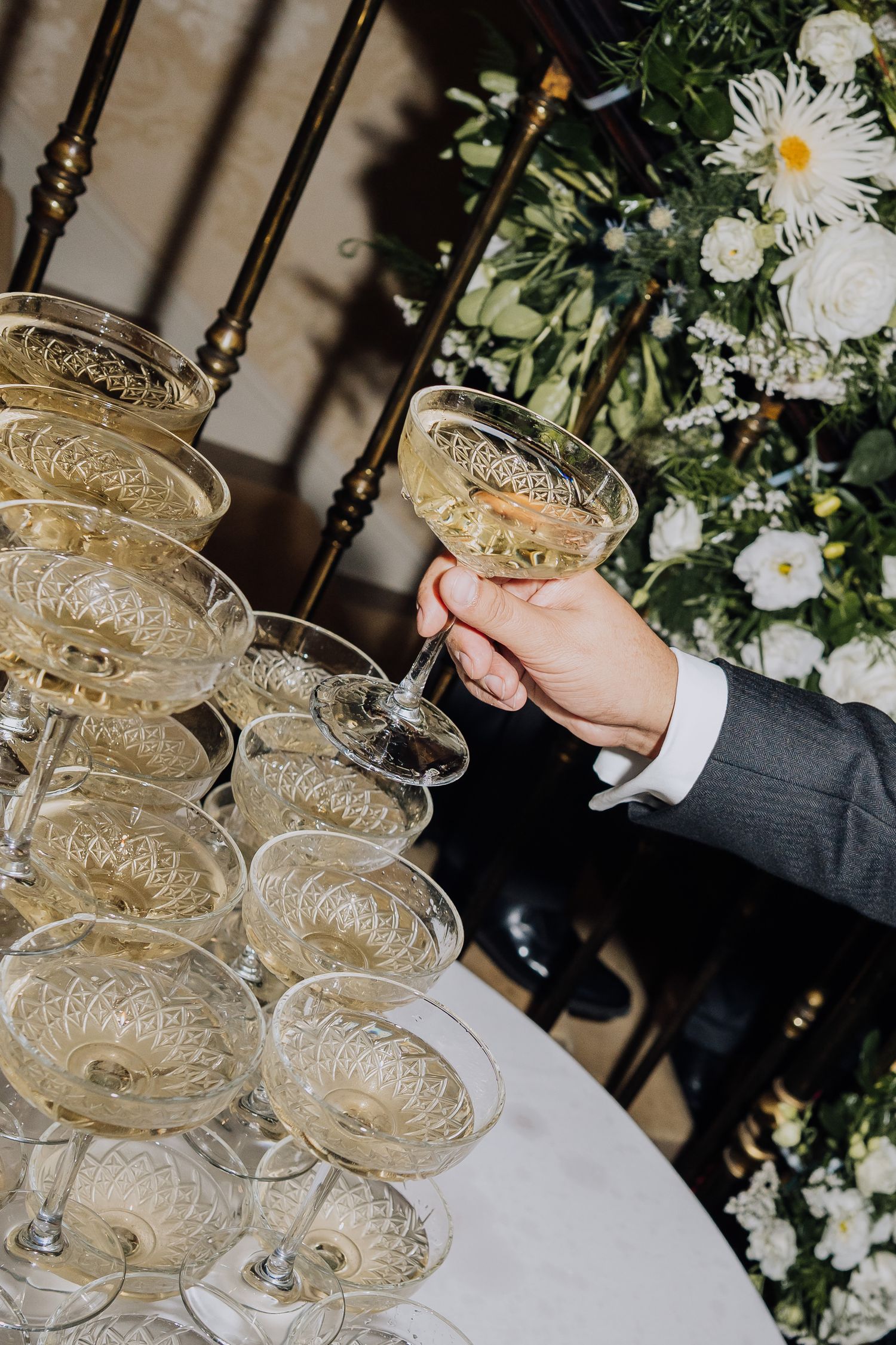 Hand taking a champagne coupe from an elegant crystal glass tower at a wedding reception with white floral backdrop.