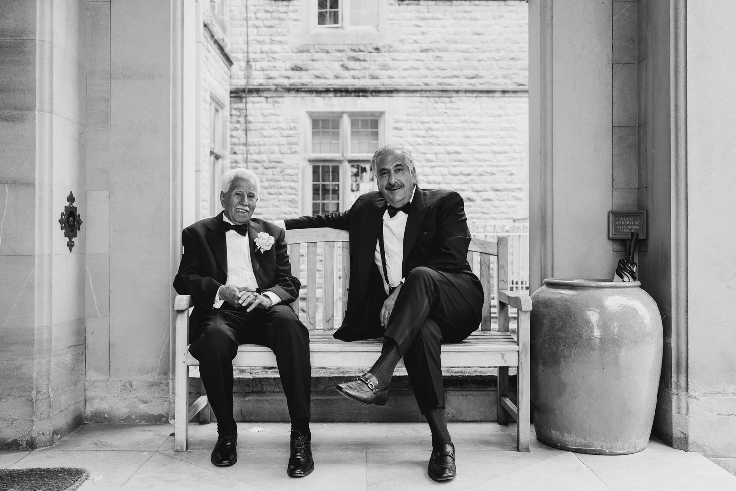 Two elderly men in tuxedos sitting on bench at elegant wedding venue in black and white.