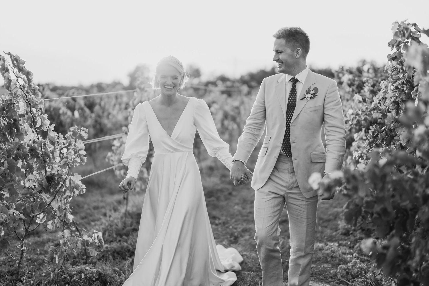 Black and white photo of smiling bride and groom holding hands while walking through a vineyard.