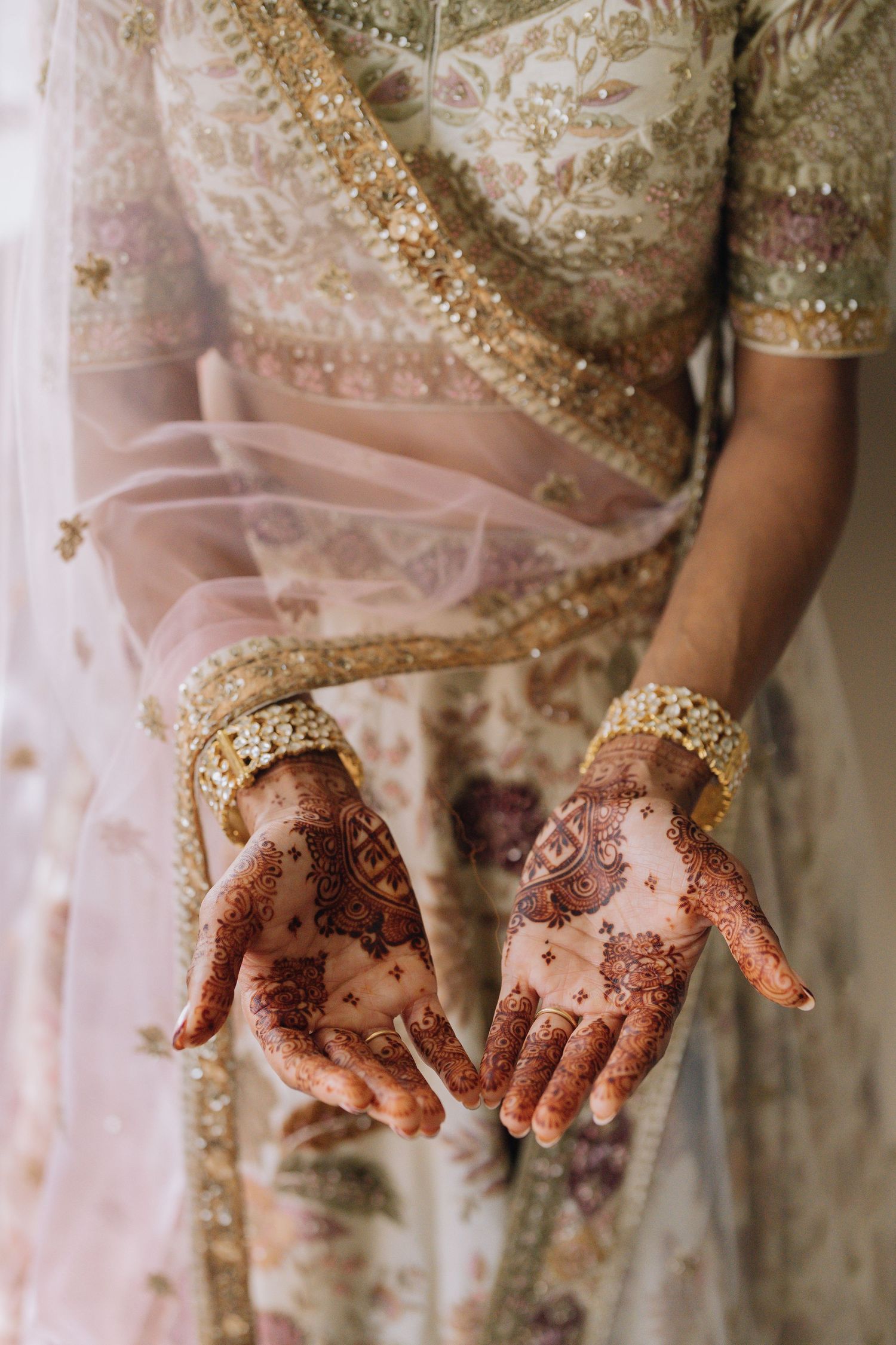 Bride displays intricate henna mehndi designs on palms, wearing gold bangles and embroidered floral lehenga.