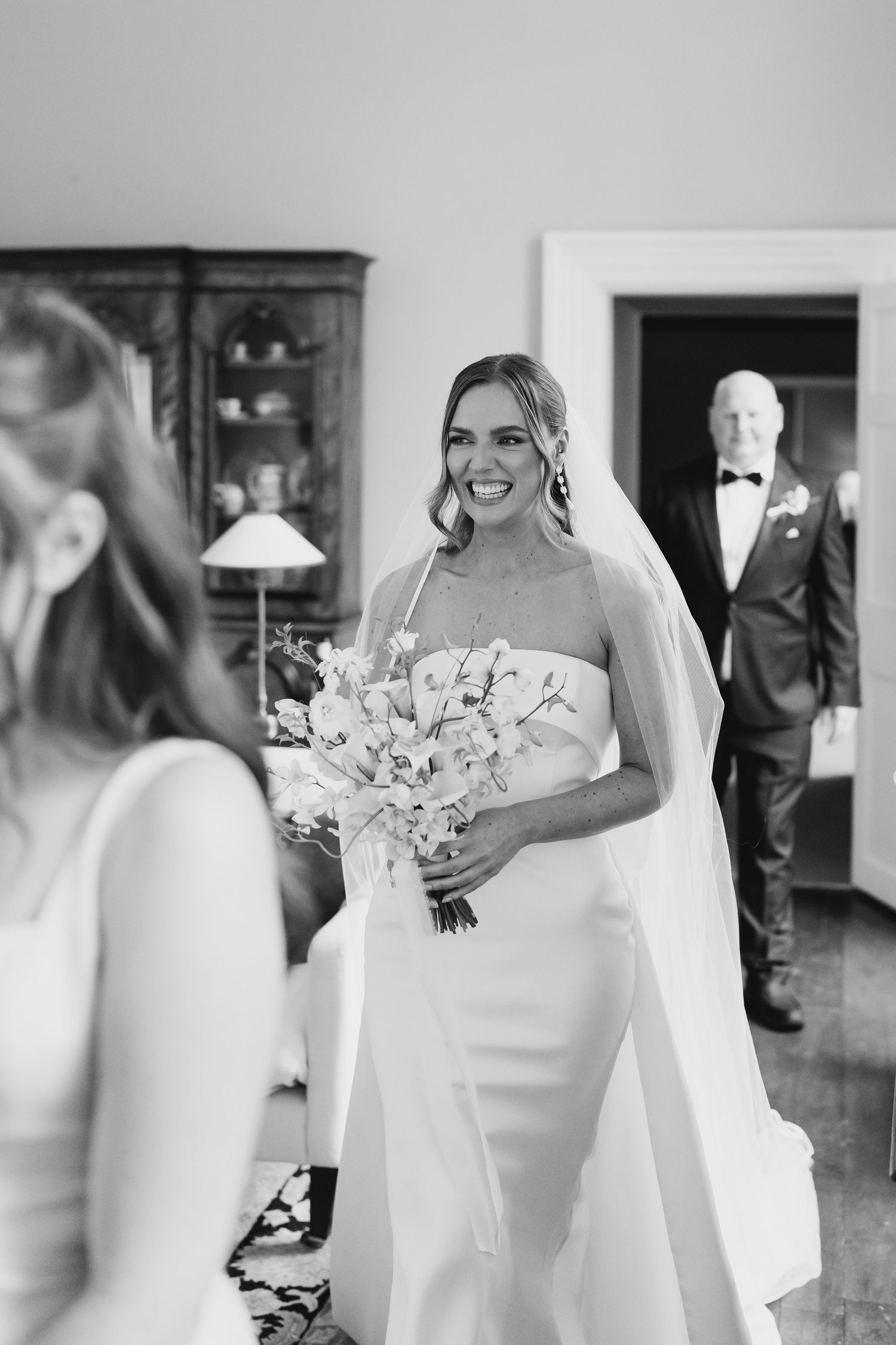 Smiling bride in strapless white gown holds bouquet while father in tuxedo stands proudly in background.