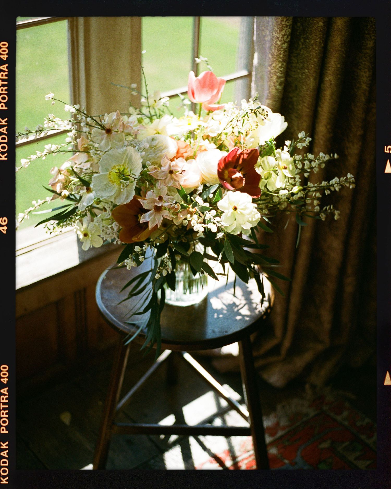 Lush floral arrangement of white, pink, and coral blooms in a vase on a dark stool by a sunlit window.
